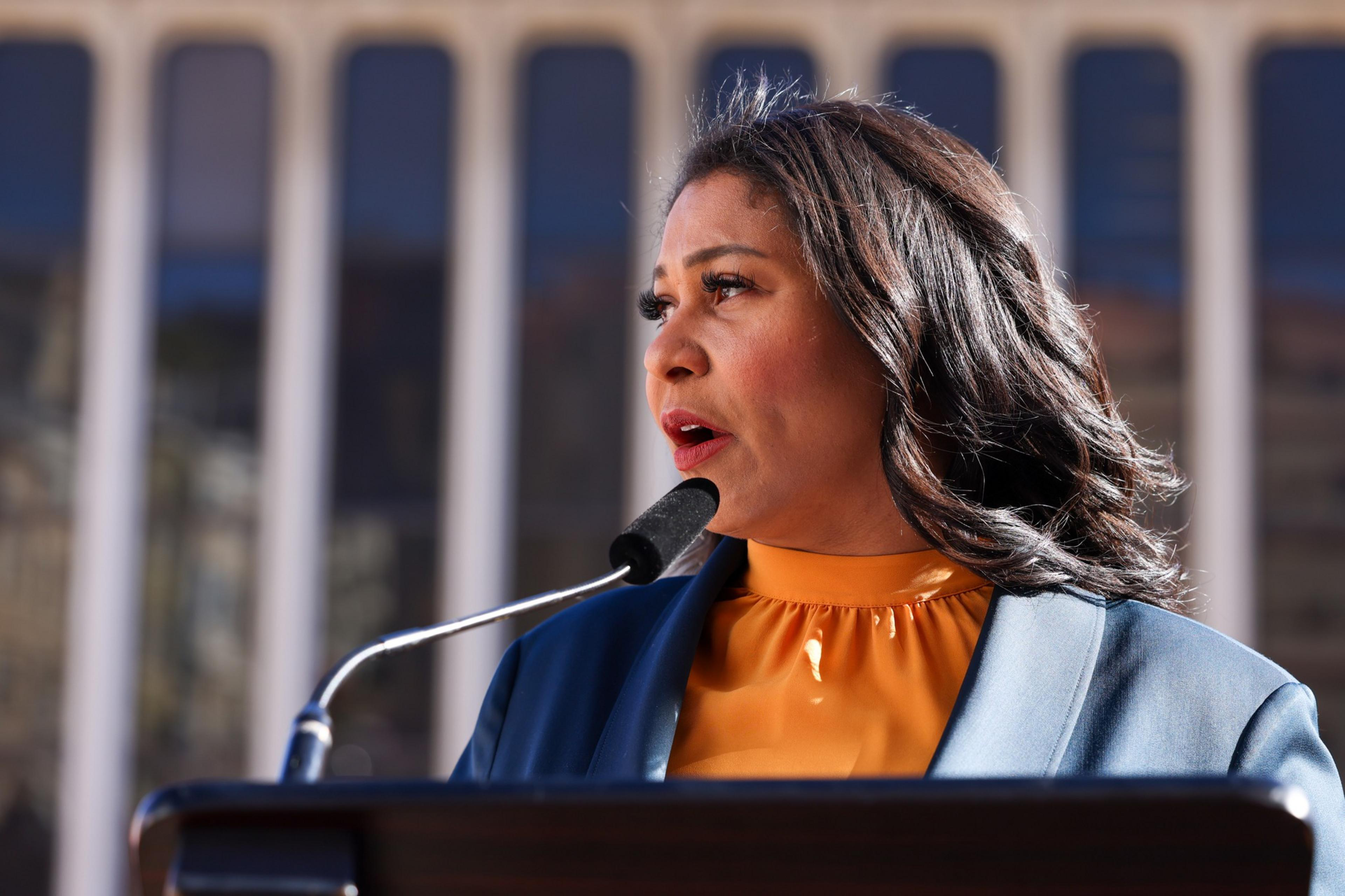 A woman with dark wavy hair speaks into a microphone at a podium, wearing an orange top and blue jacket, in front of a modern building with tall, vertical windows.