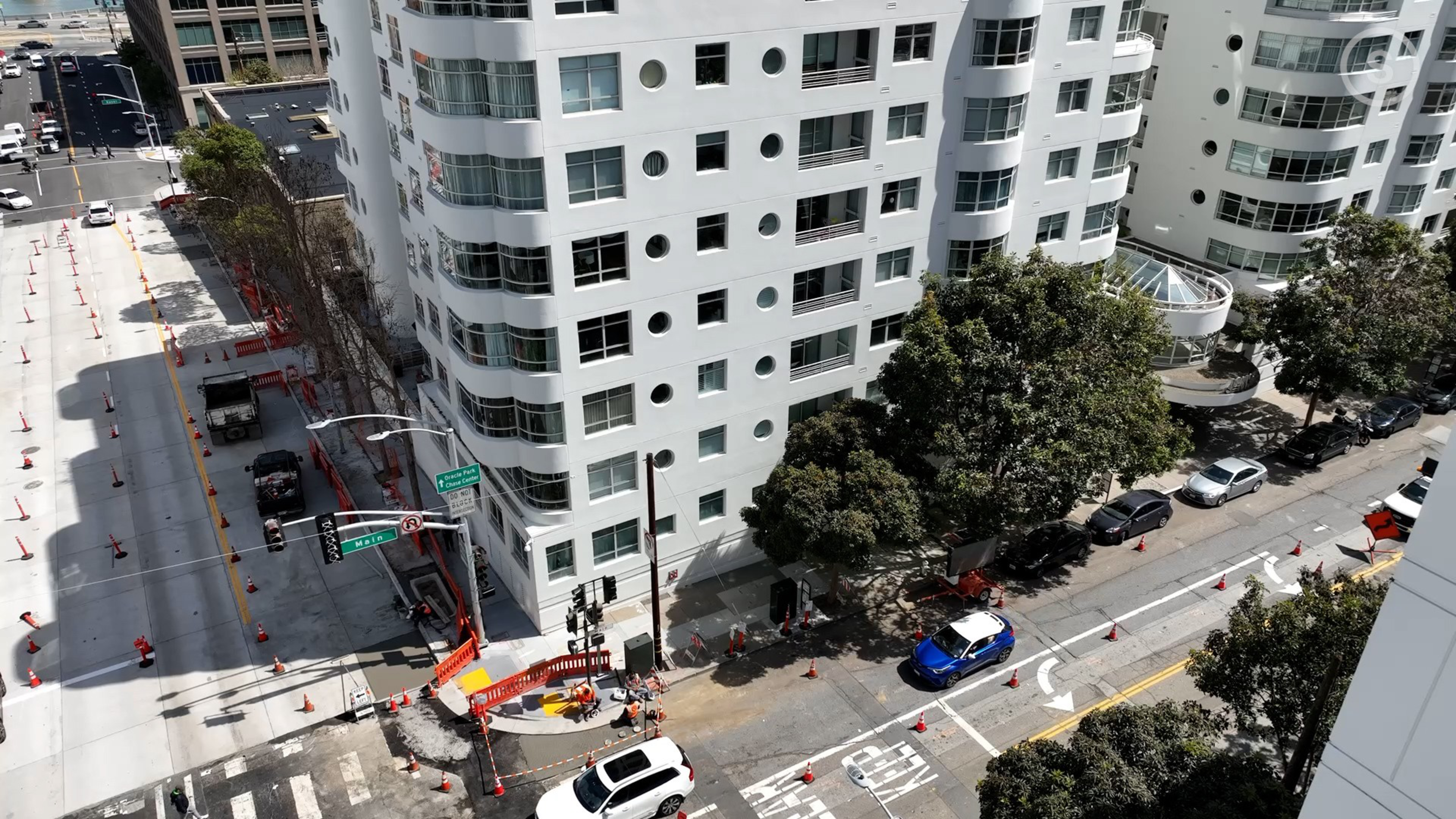A street corner with multi-story white buildings, a construction zone with orange cones, several parked cars, and trees lining the sidewalk.