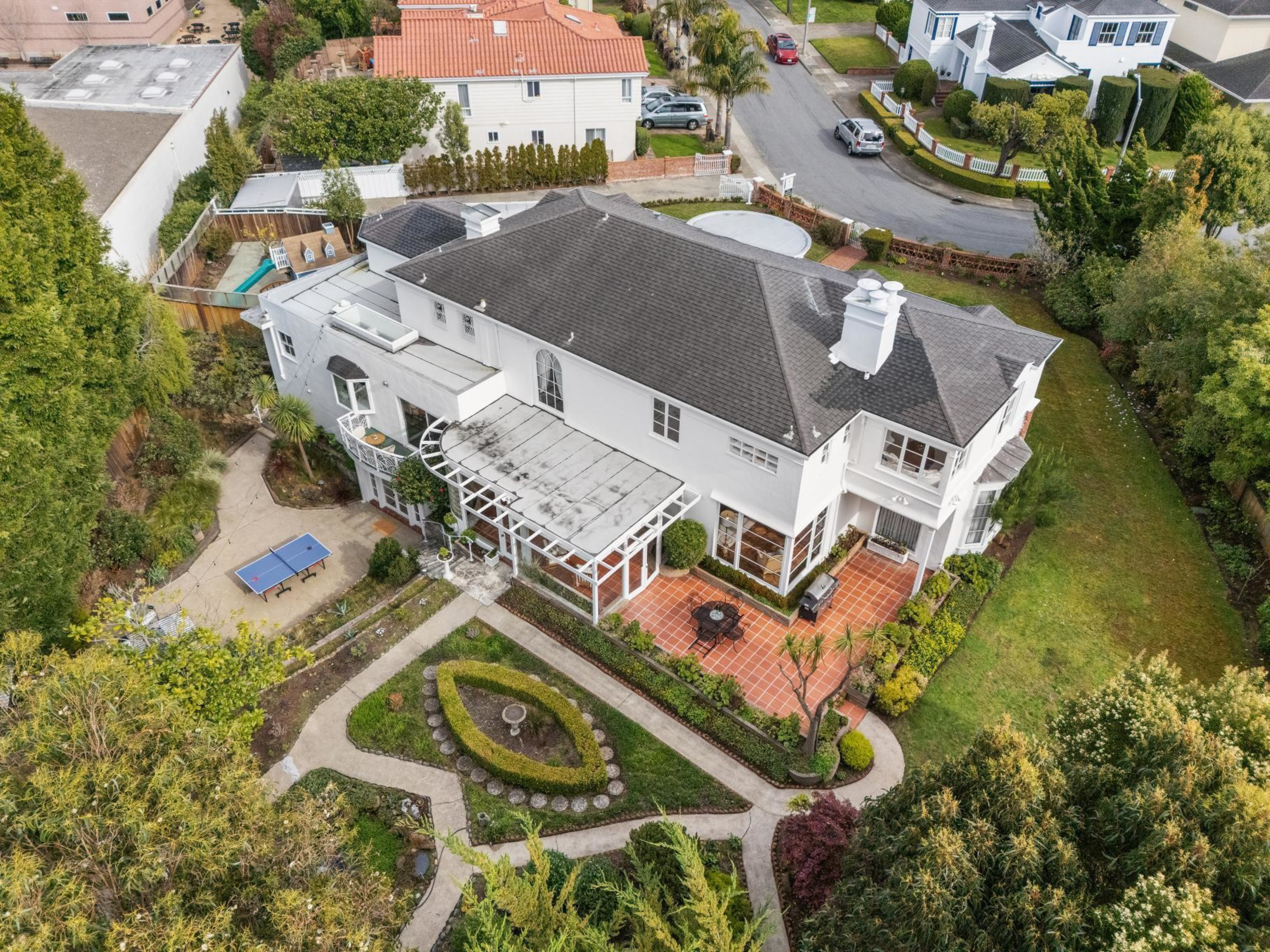 A large white two-story house with a gray roof, surrounded by manicured gardens, patio furniture, a pergola, a ping pong table, and adjacent streets.