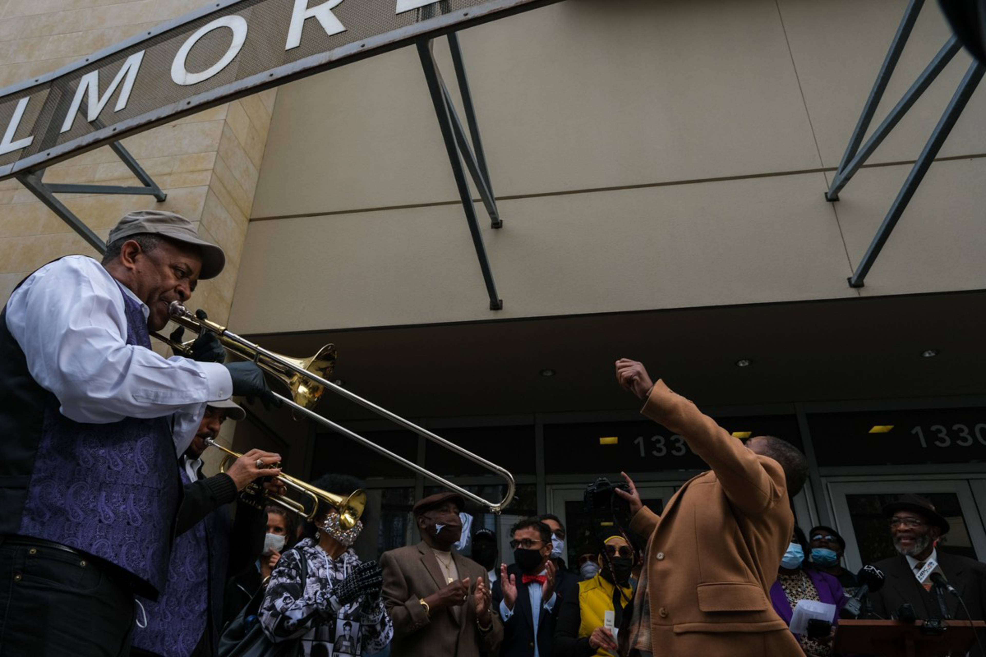 A street band with trombone and trumpet plays while a lively crowd claps and a man in a brown jacket raises his arm in excitement.