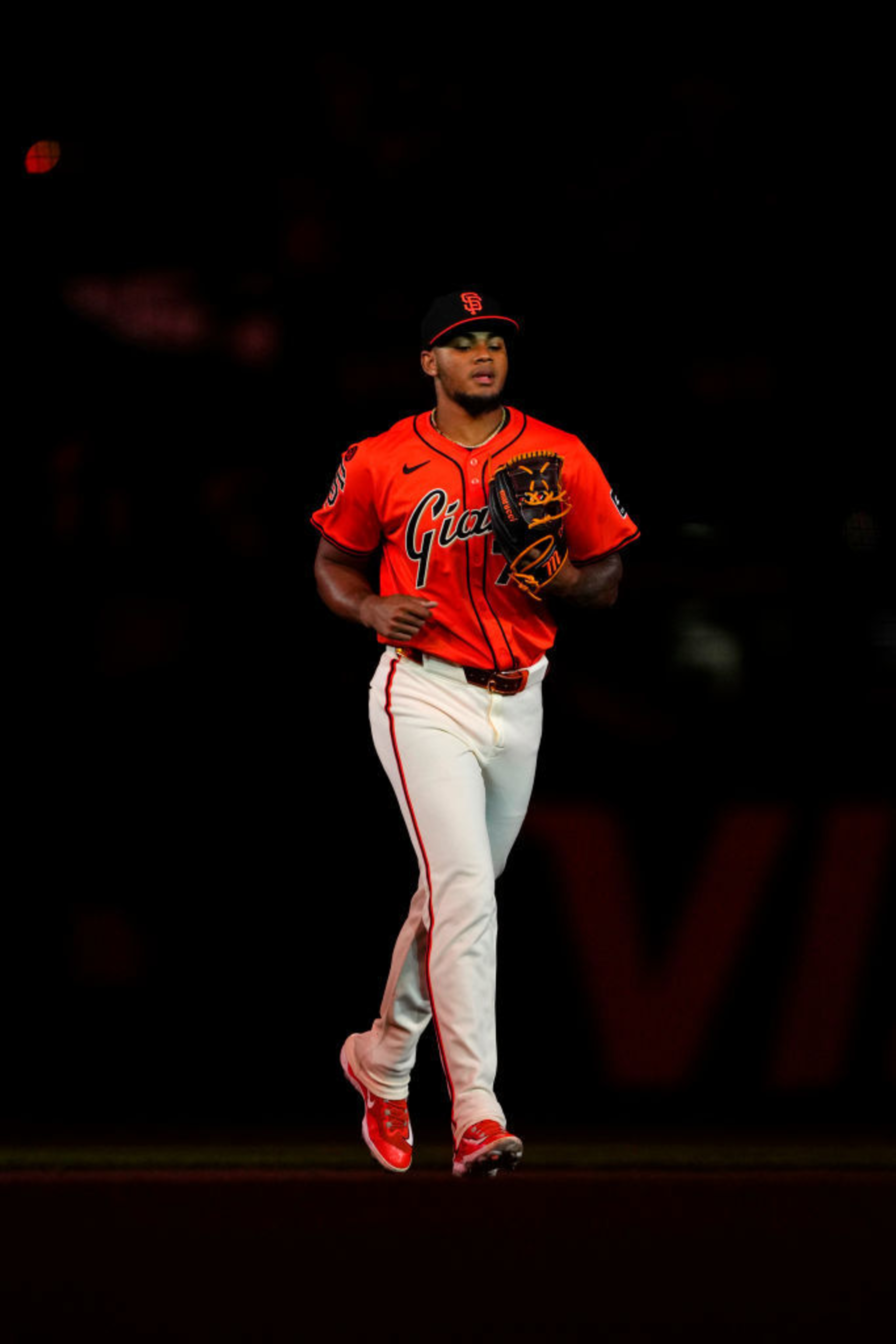 A baseball player is running on the field, wearing an orange jersey and white pants. The jersey reads &quot;Giants,&quot; and he holds a glove with the team logo on his cap.