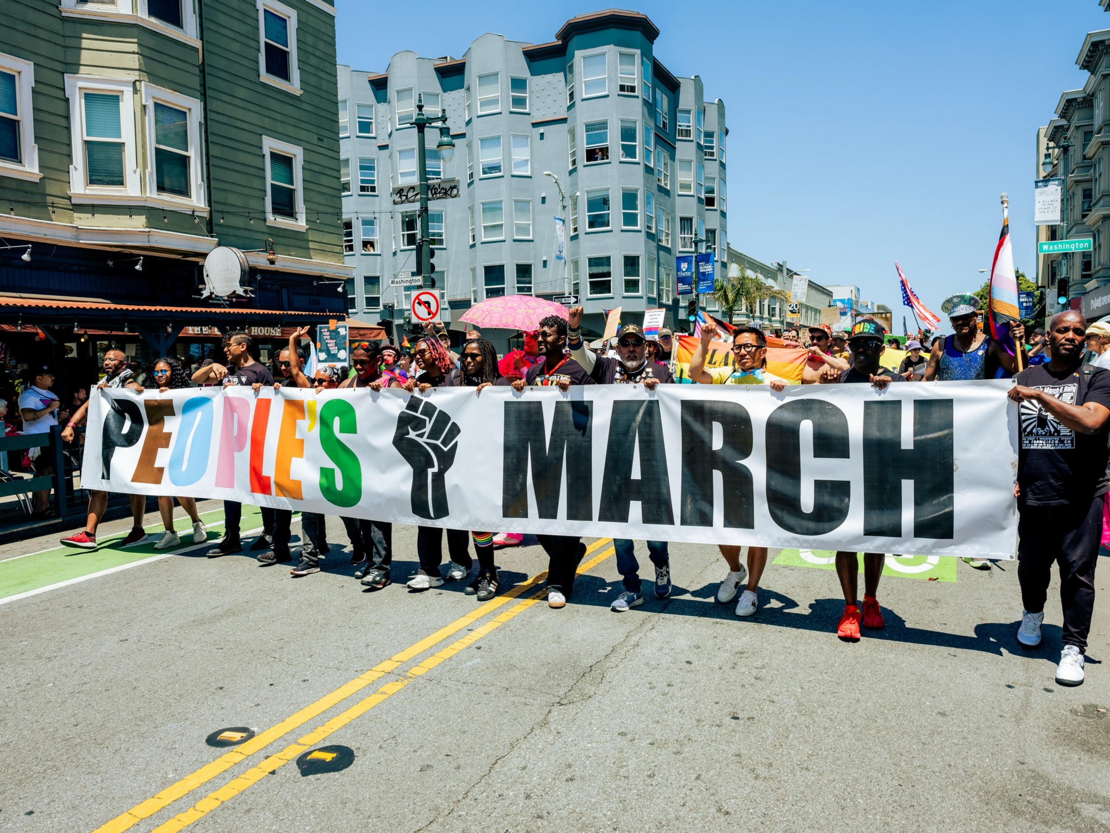A group of people are walking in a street, holding a large banner that says "PEOPLE'S MARCH" with a raised fist graphic. Colorful buildings and flags are in the background.