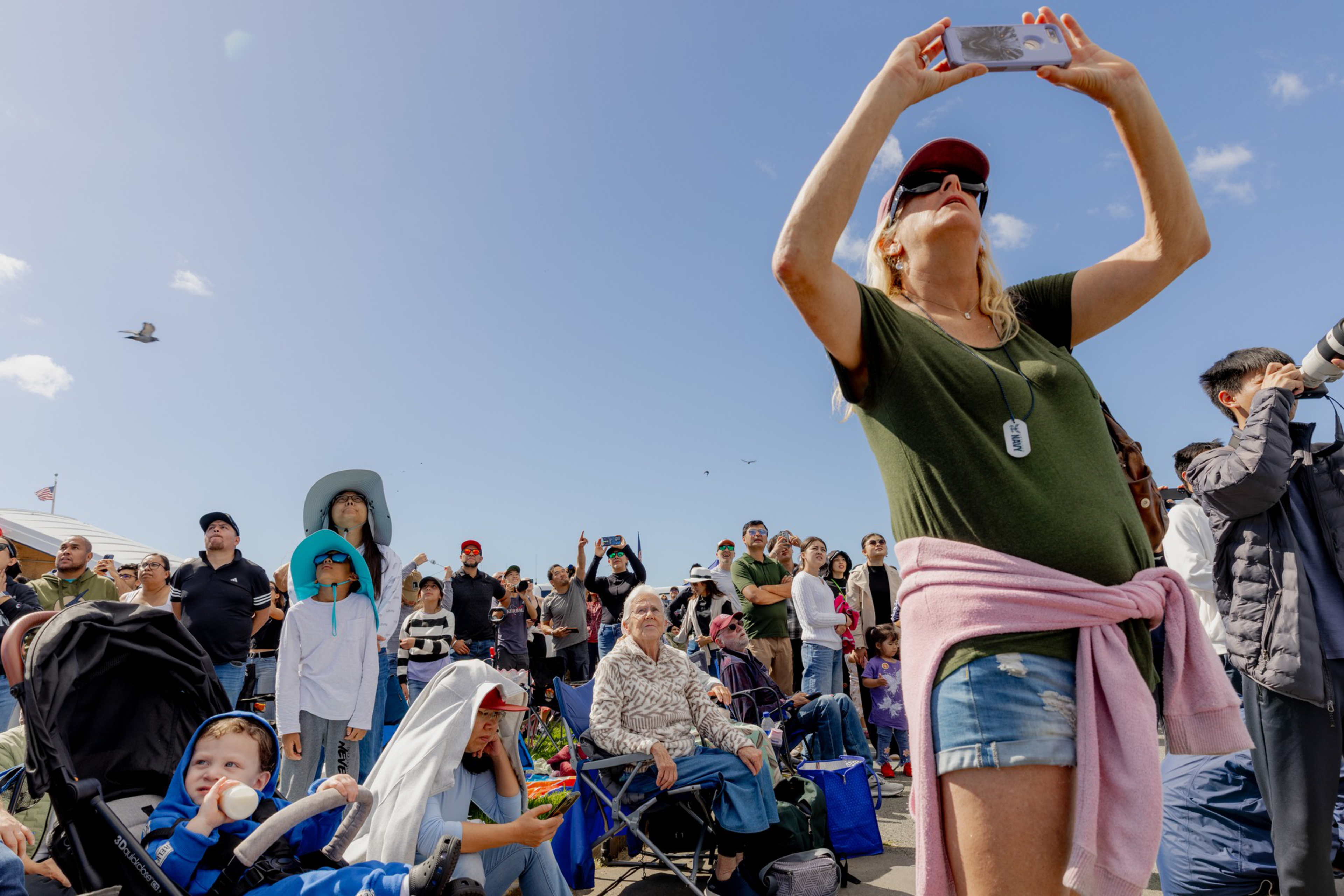 A diverse crowd of people, including children and adults, looks skyward on a sunny day as some take photos and others watch intently.