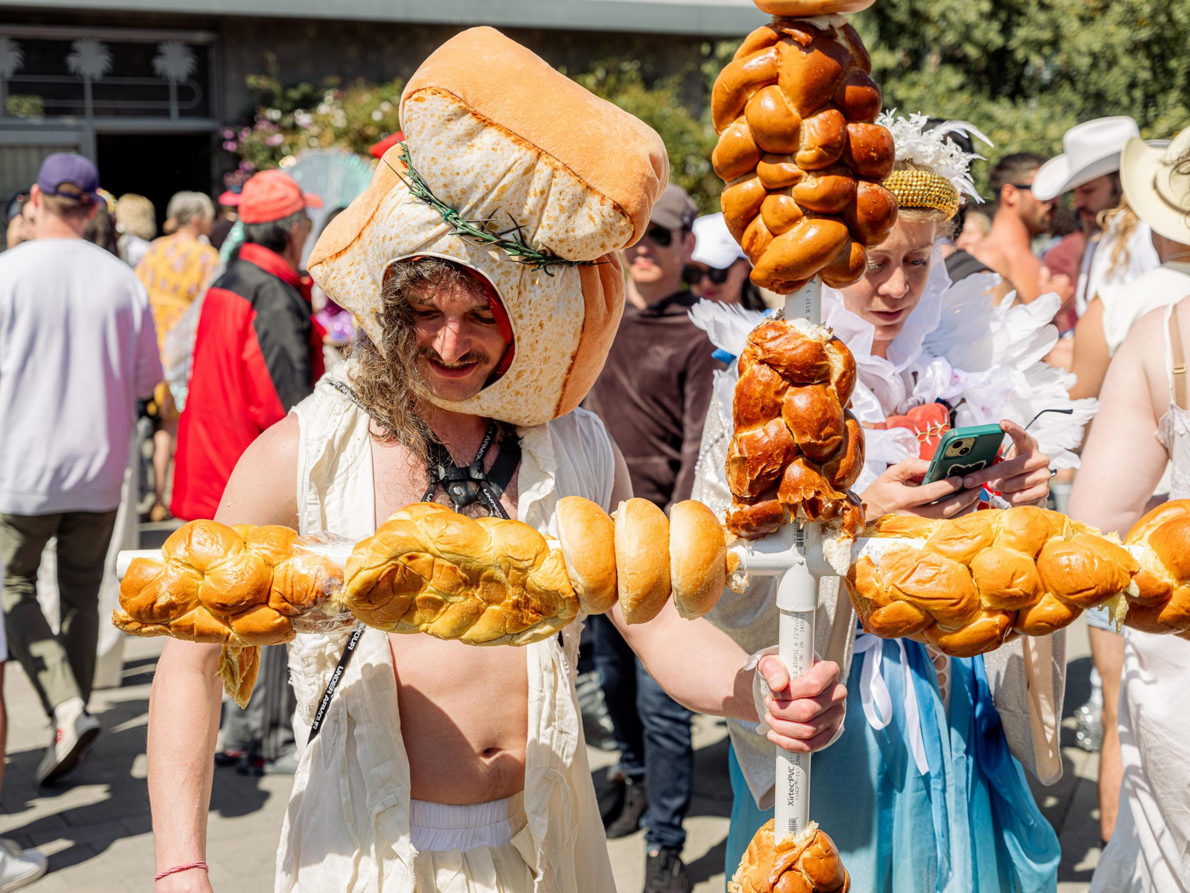 A person wears a loaf-shaped hat and holds a pole adorned with various breads, surrounded by a crowd. Another person looks at a phone beside them.