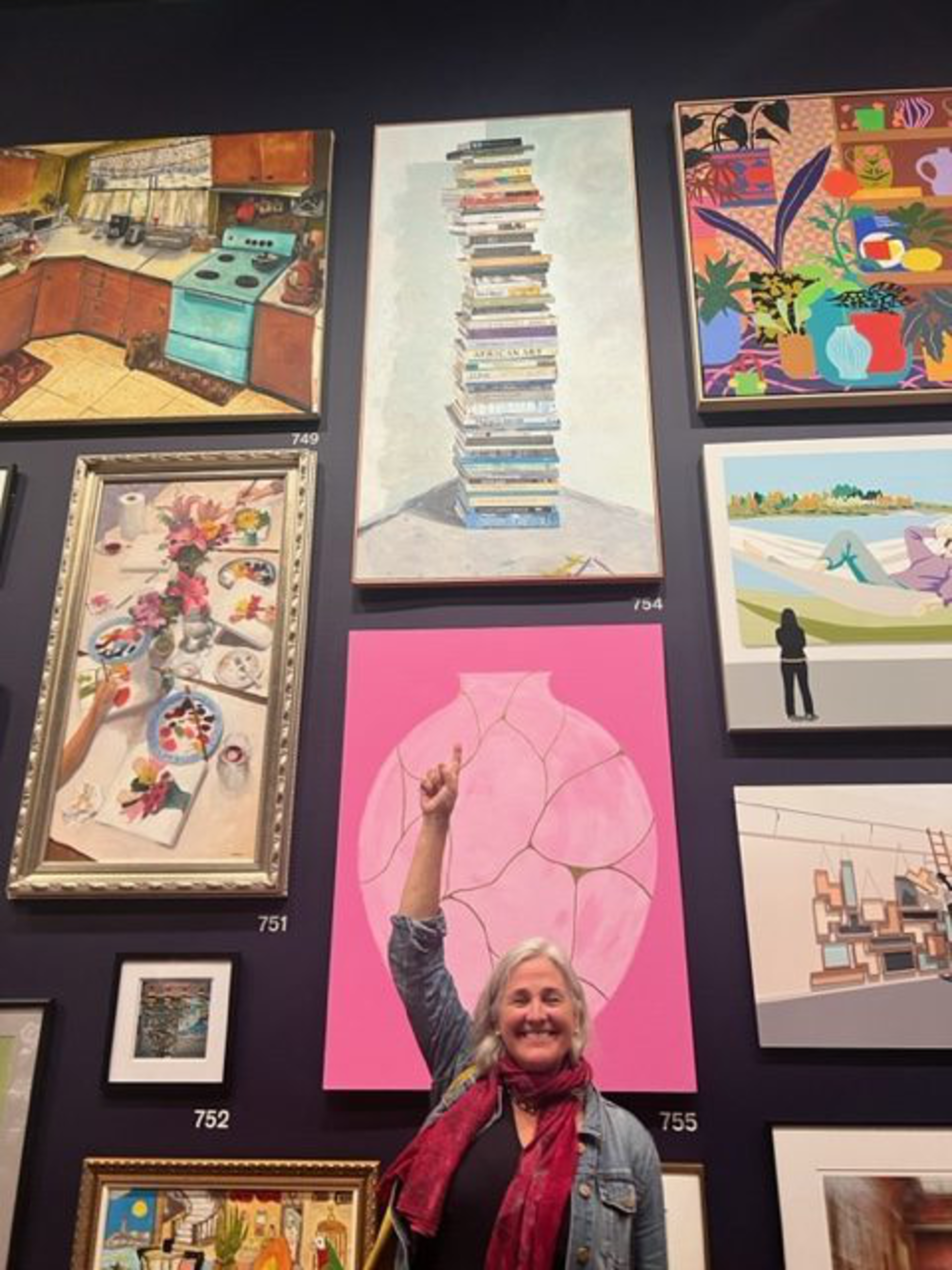 A woman smiles and points upward toward a gallery wall displaying various paintings, including a kitchen scene, a tall stack of books, and a large pink vase.