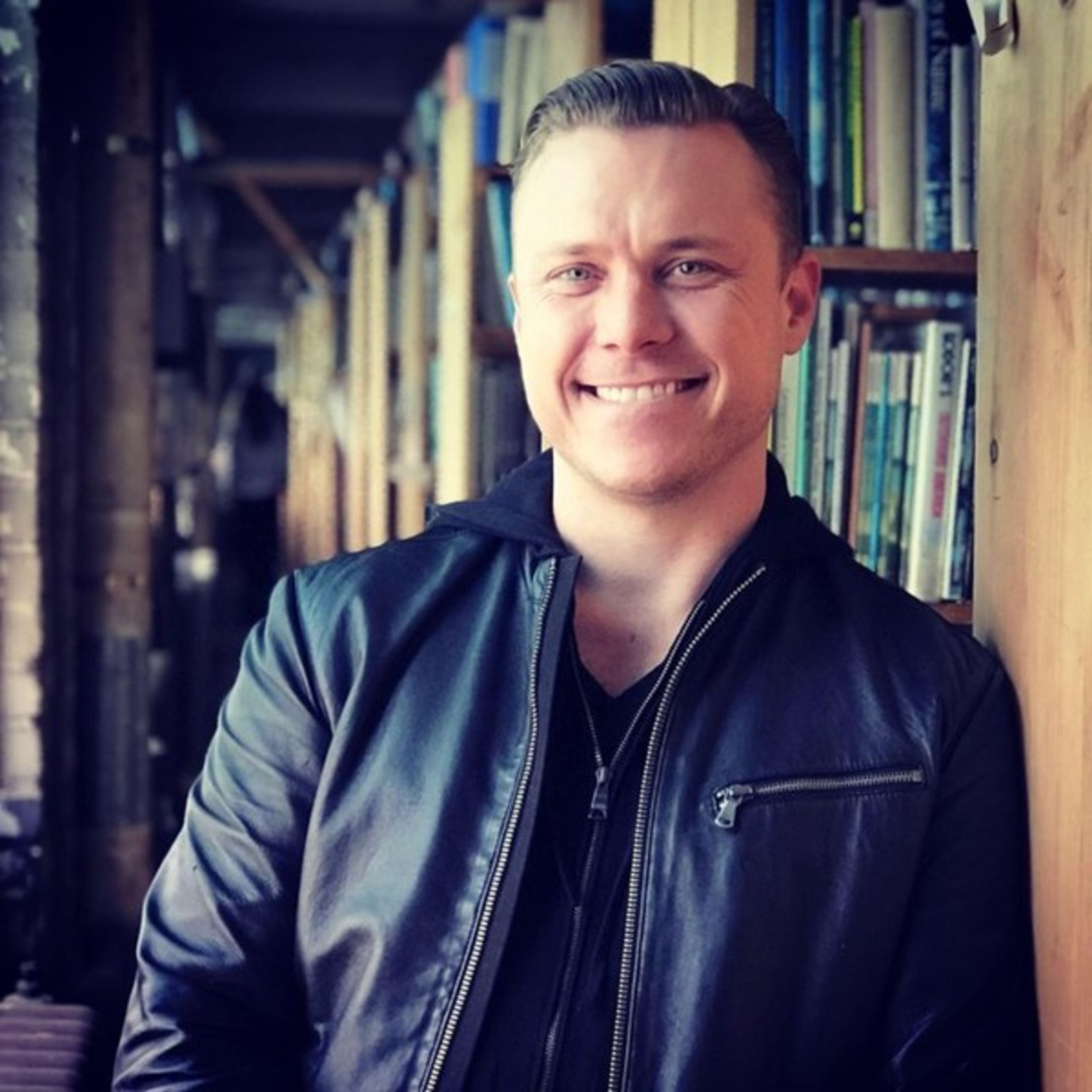 A smiling man in a black jacket stands next to bookshelves in a warmly lit room. The background is filled with books and soft shadows.