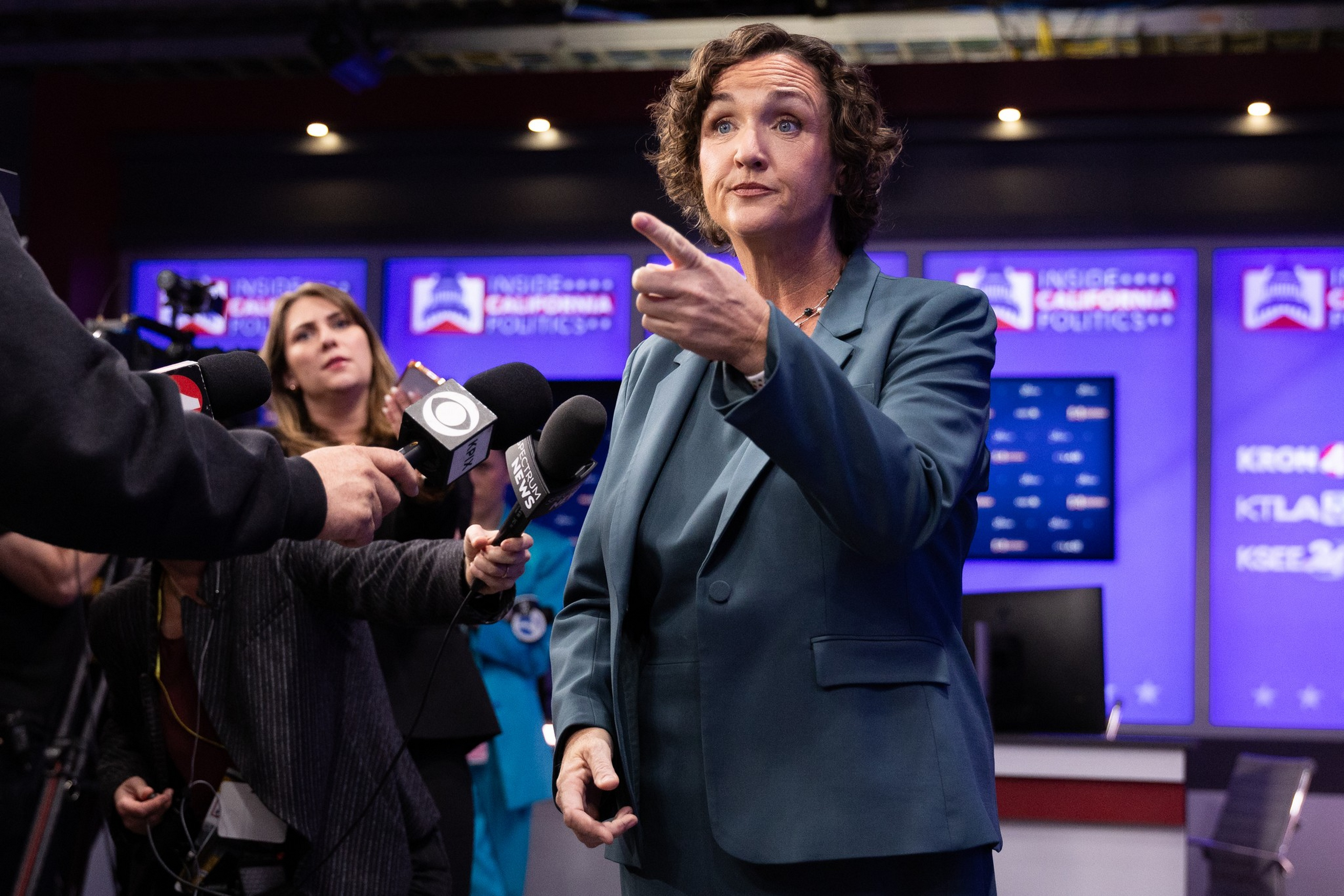 A woman in a blue suit points while speaking to reporters holding microphones during a political event with screens in the background.