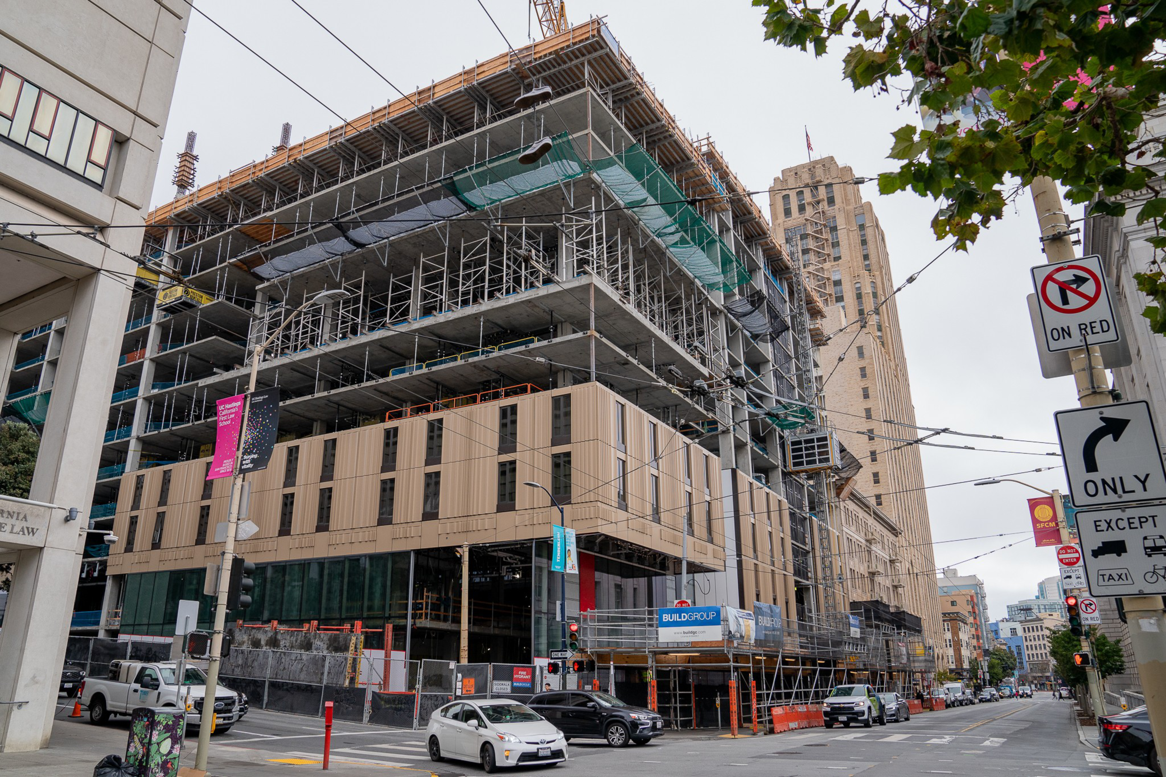 A large multi-story building under construction stands at a street corner with scaffolding, construction signs, traffic lights, and vehicles nearby.