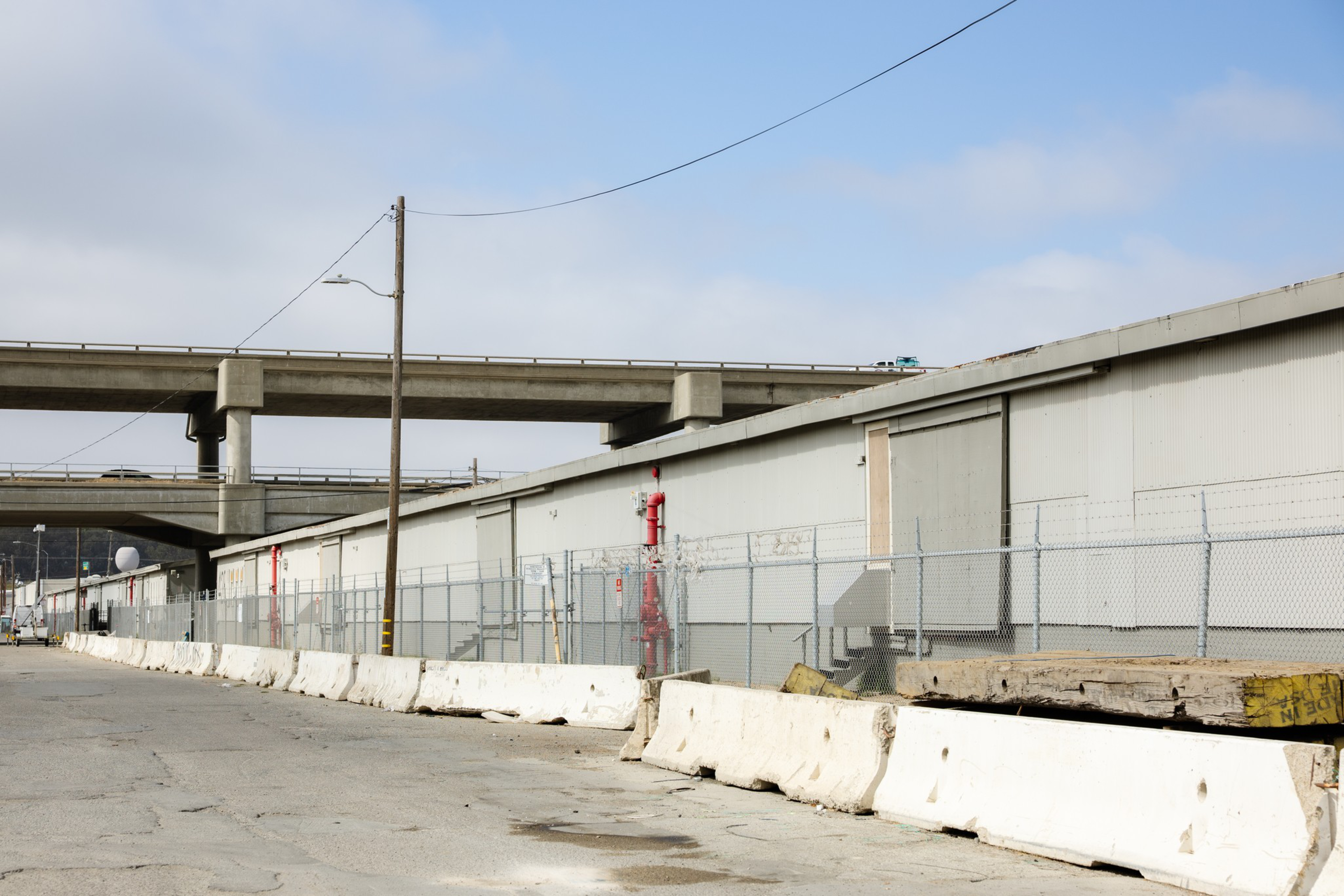 A long gray warehouse with chain-link fencing in front and white concrete barriers lining a cracked, empty street under a partly cloudy sky.