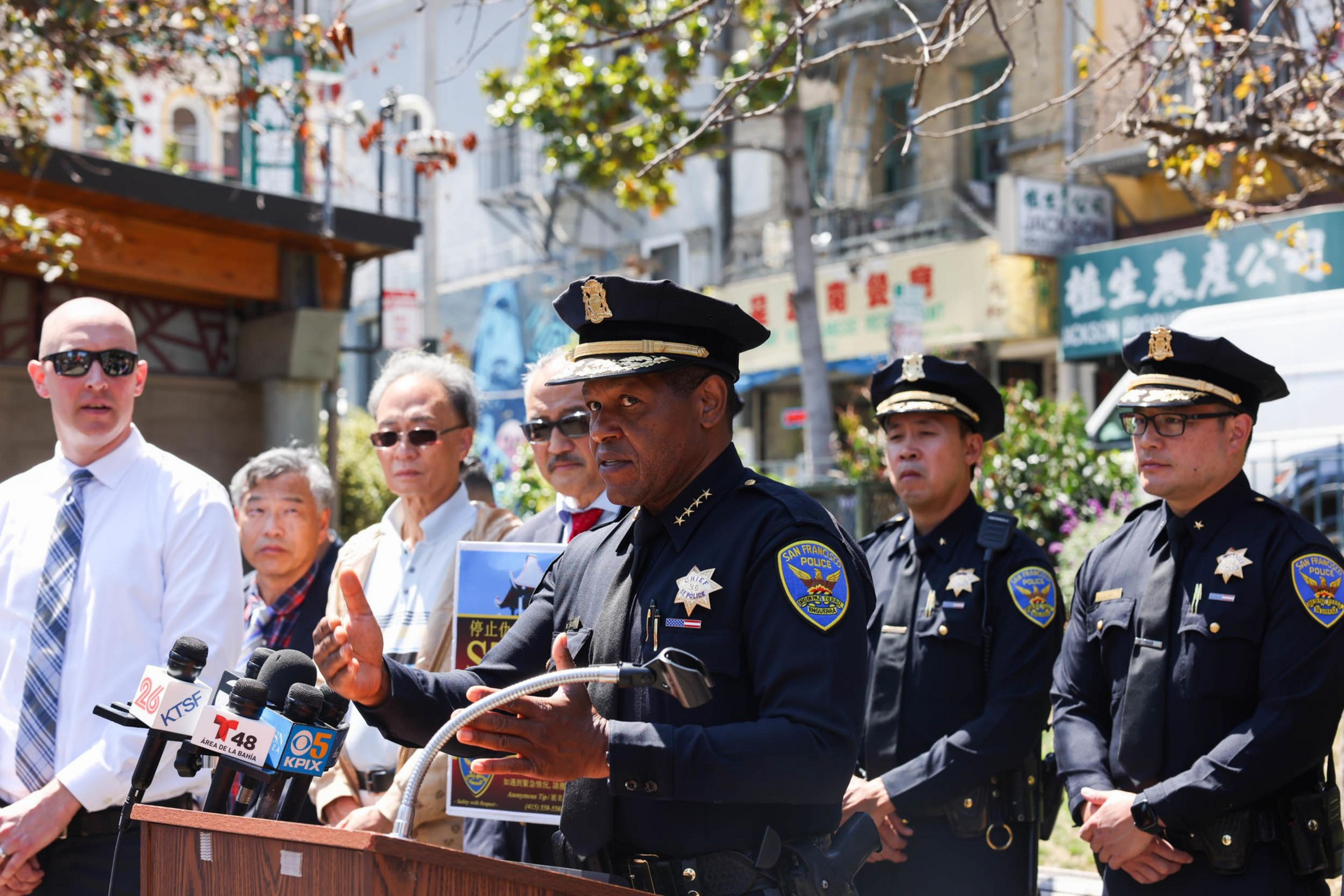 A police officer speaks at a podium with microphones while five men, including three other uniformed officers, listen behind him outdoors.