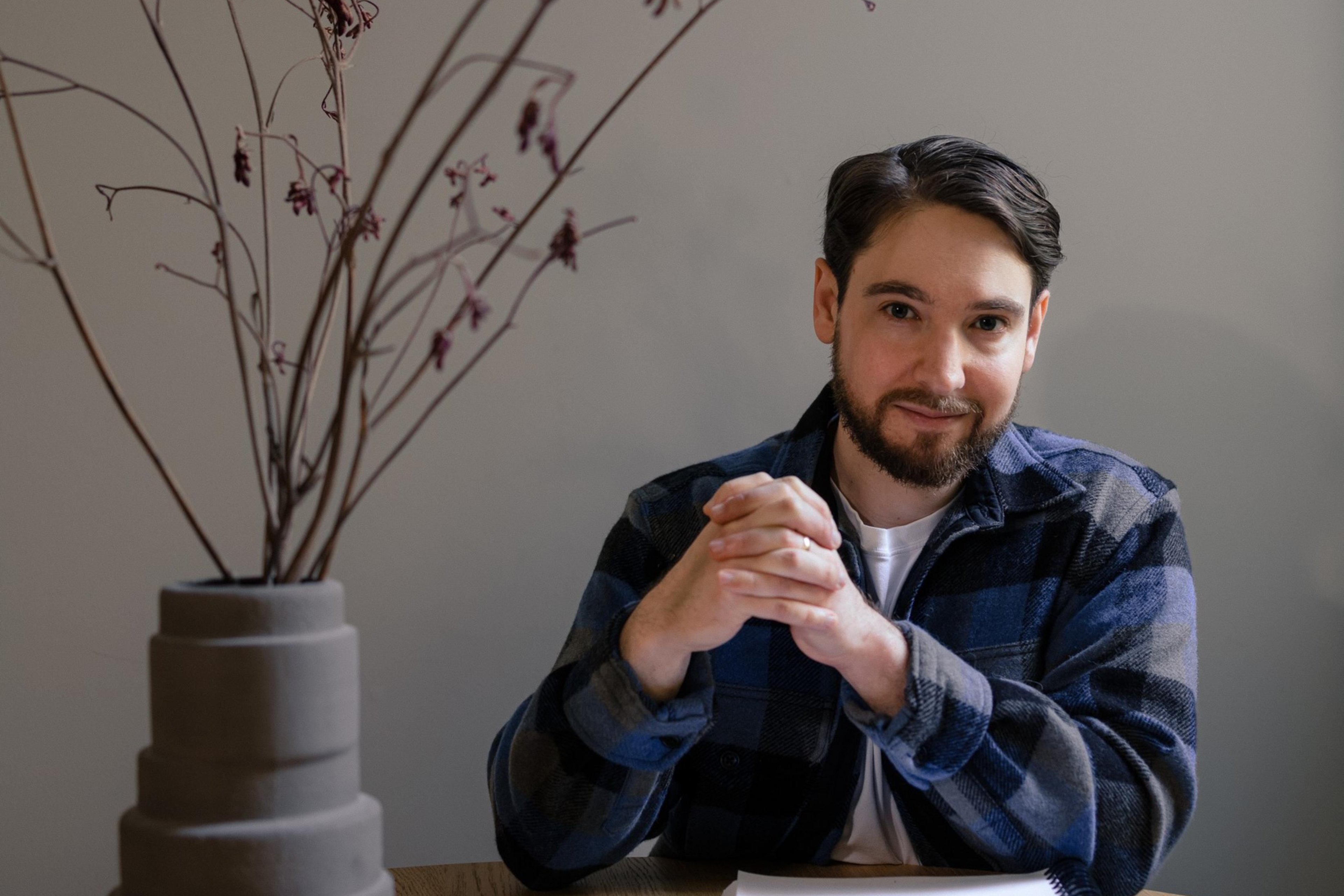 A man with dark hair and a beard sits at a table, hands clasped, wearing a blue plaid shirt. A tall vase with twigs is beside him, and a notepad is in front.