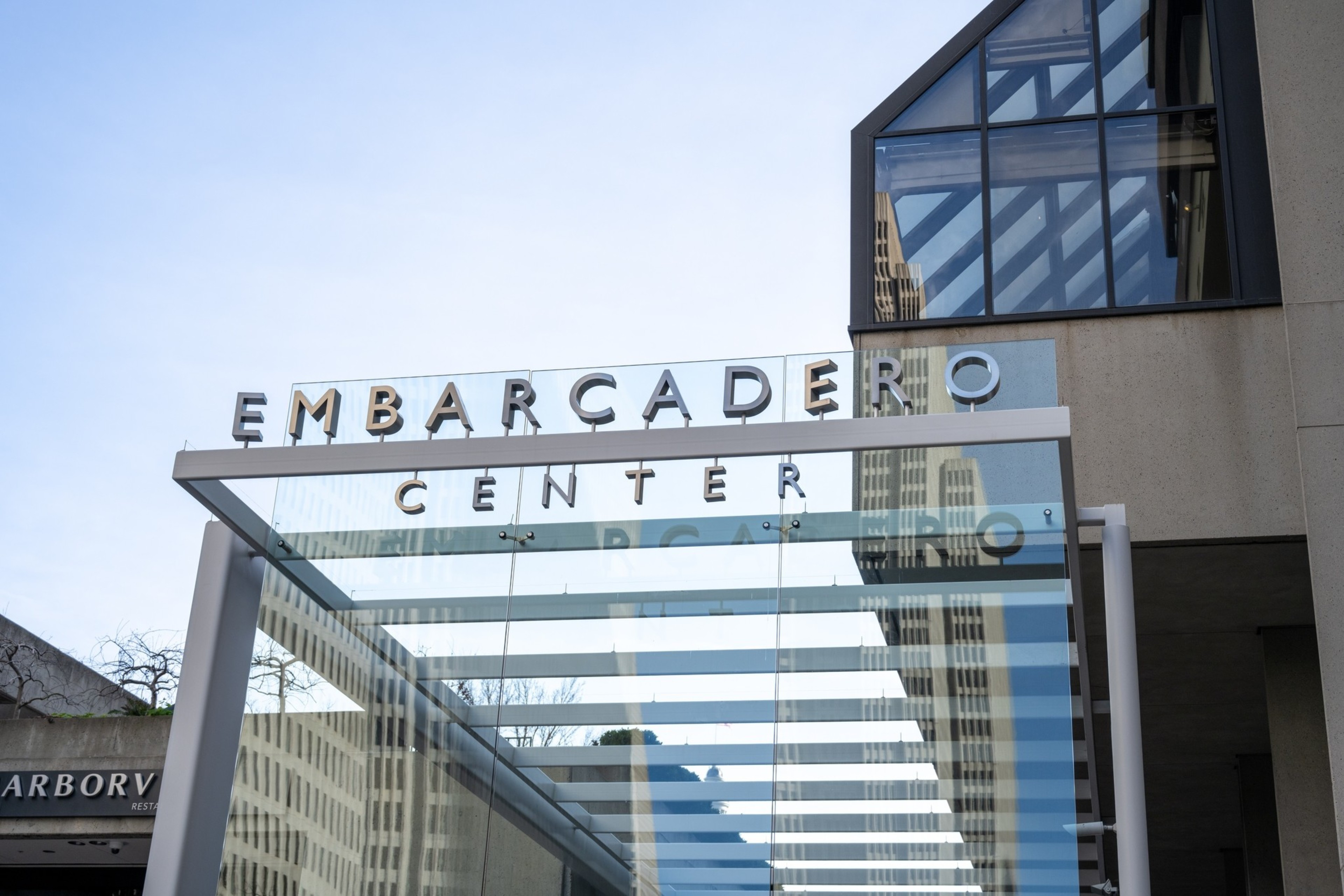 The image shows a modern glass entrance with &quot;EMBARCADERO CENTER&quot; written in bold letters above it against a clear blue sky, with a nearby building in view.