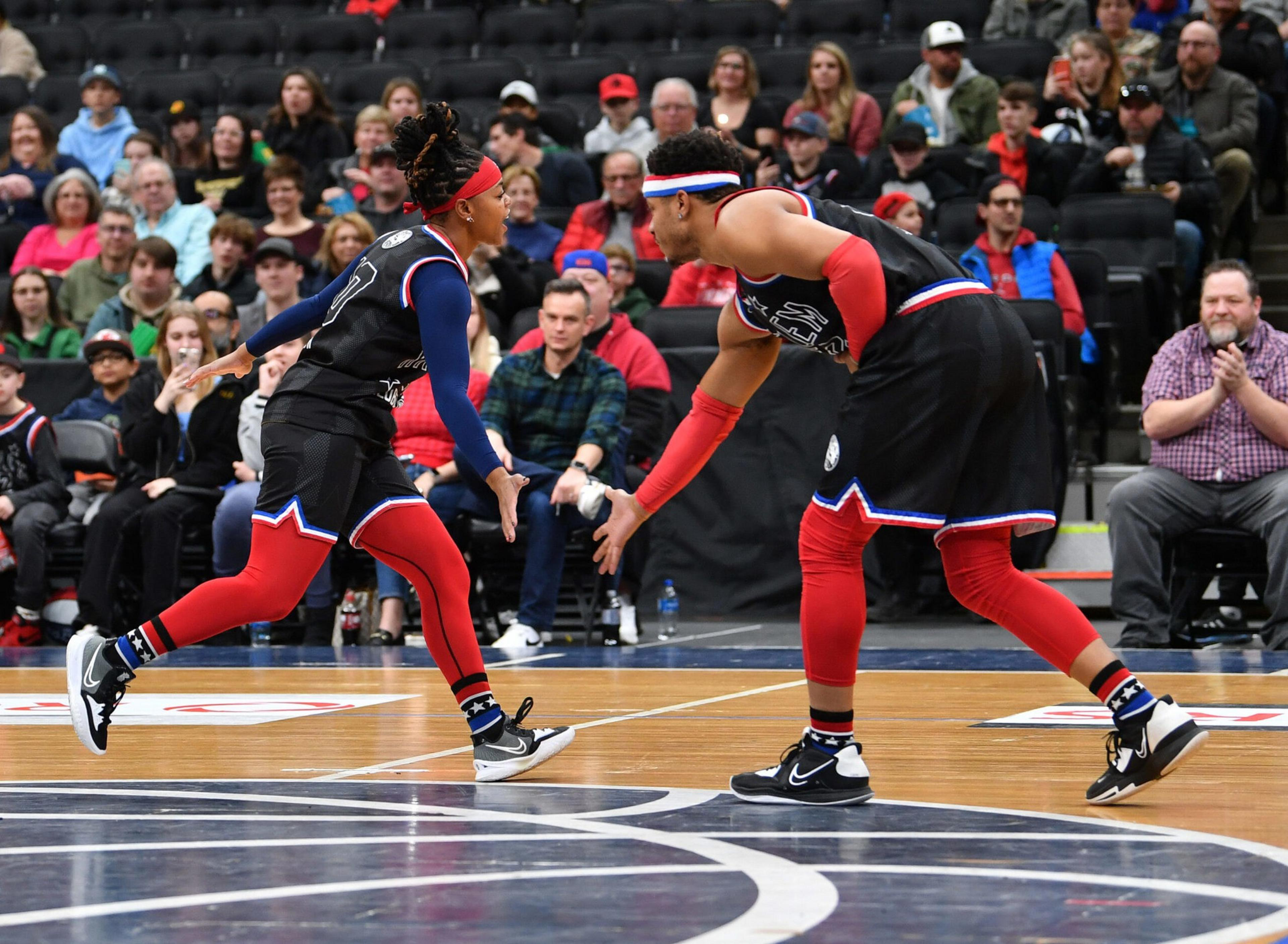 Two basketball players in black and red uniforms reach to high-five each other on the court, with spectators watching from the stands behind them.