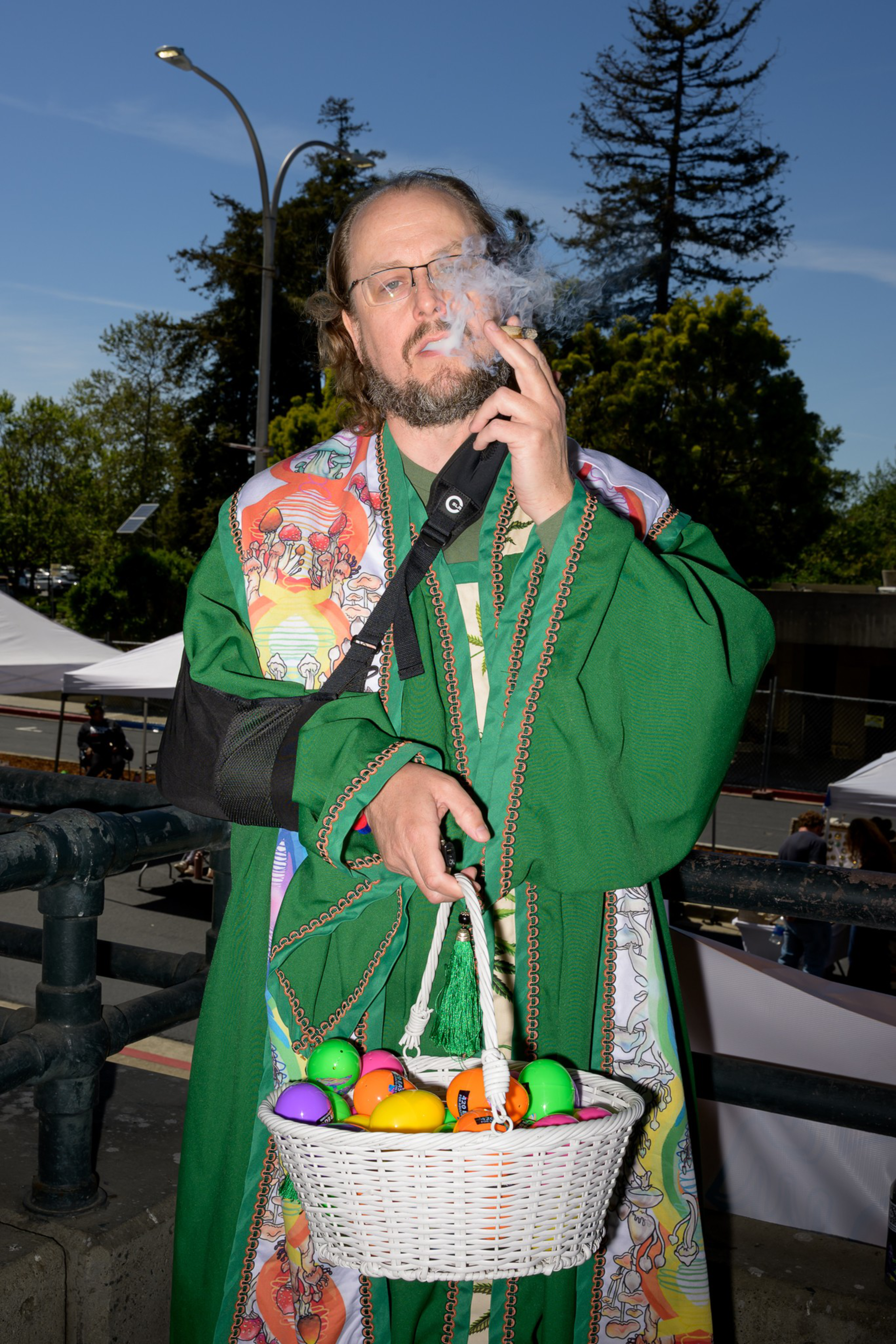 A person in a green robe and colorful shawl smokes a cigar while holding a white basket of colored eggs. They're outside on a sunny day.