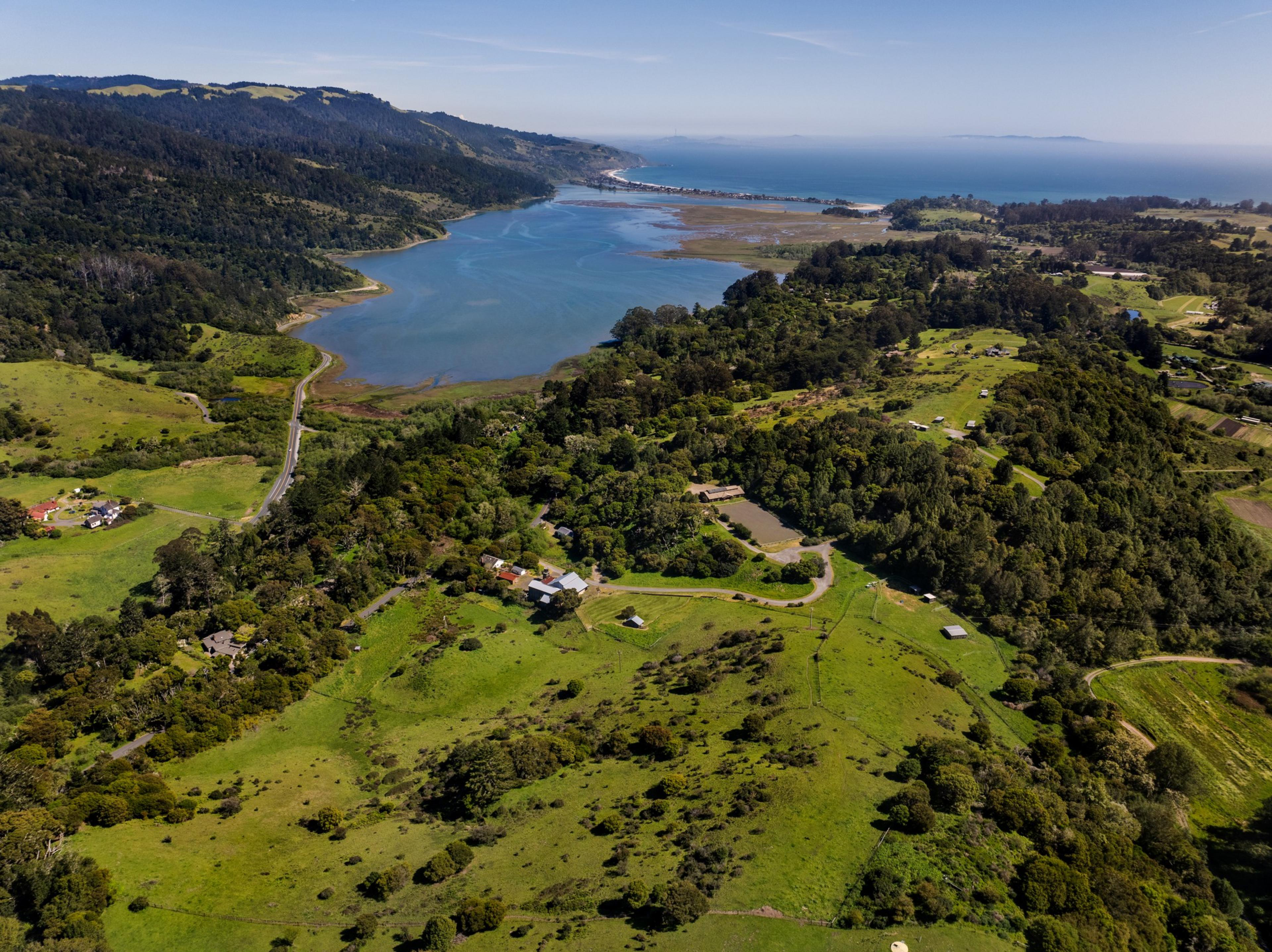 Aerial view of a lush valley with a winding river leading to the sea, dotted with small buildings and roads.