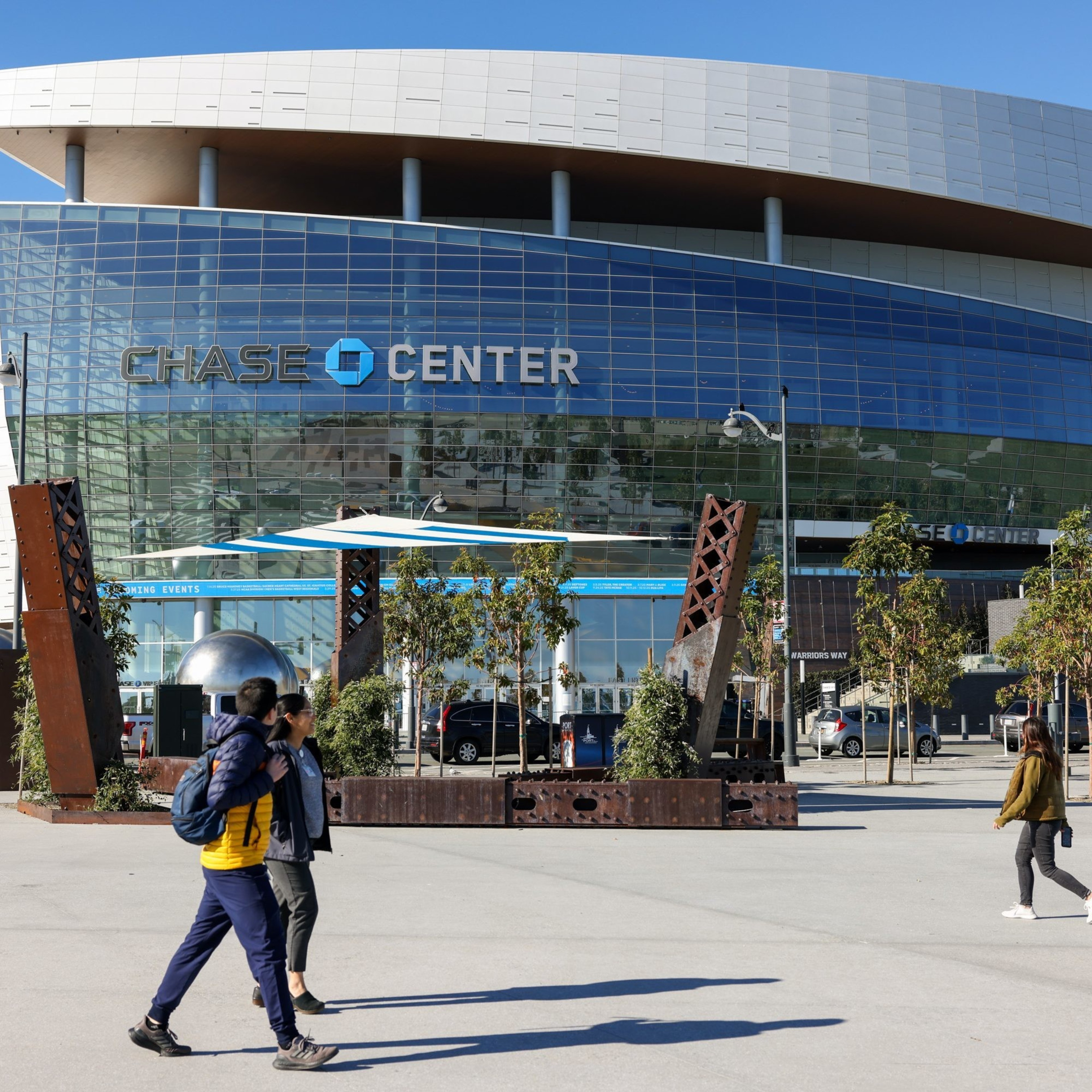 The image shows the Chase Center's modern exterior with a glass facade. People stroll in the foreground, and decorative metal sculptures and trees are present.