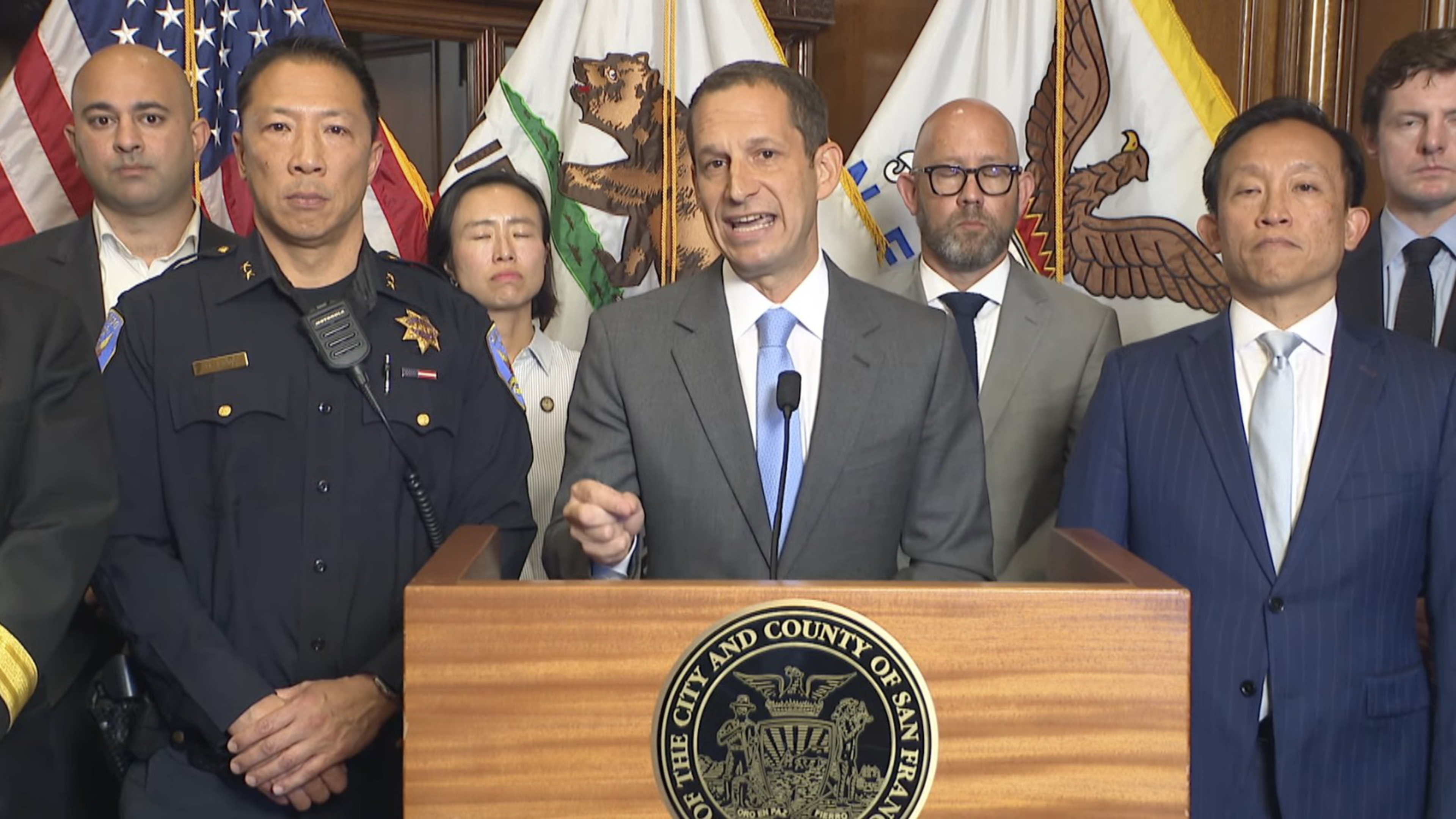 A man in a gray suit speaks at a San Francisco podium, flanked by officials including a uniformed police officer and others in suits.
