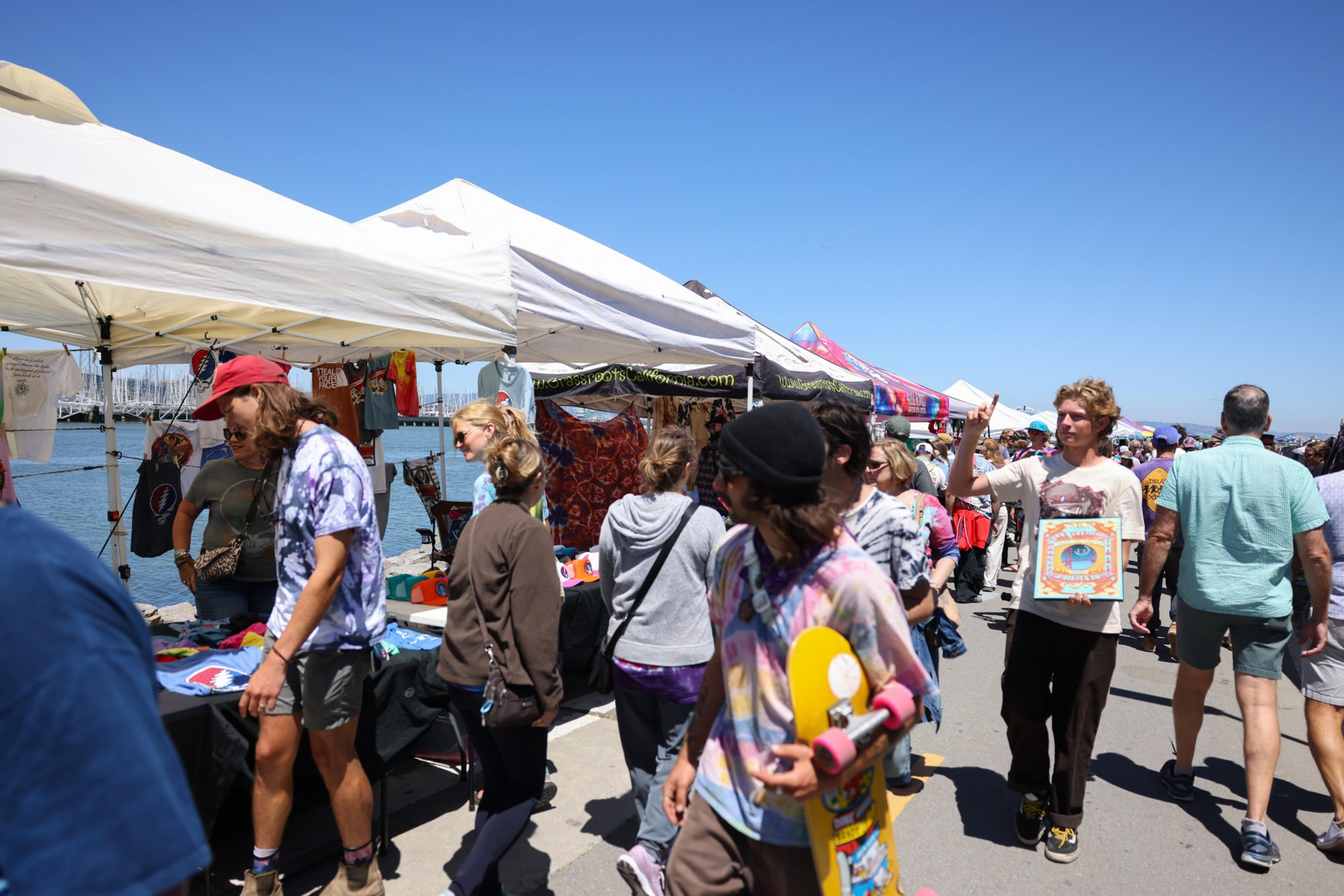 The image shows a bustling outdoor market by the waterfront with people browsing colorful stalls under tents. One person holds a skateboard, and another lifts a hand in gesture.