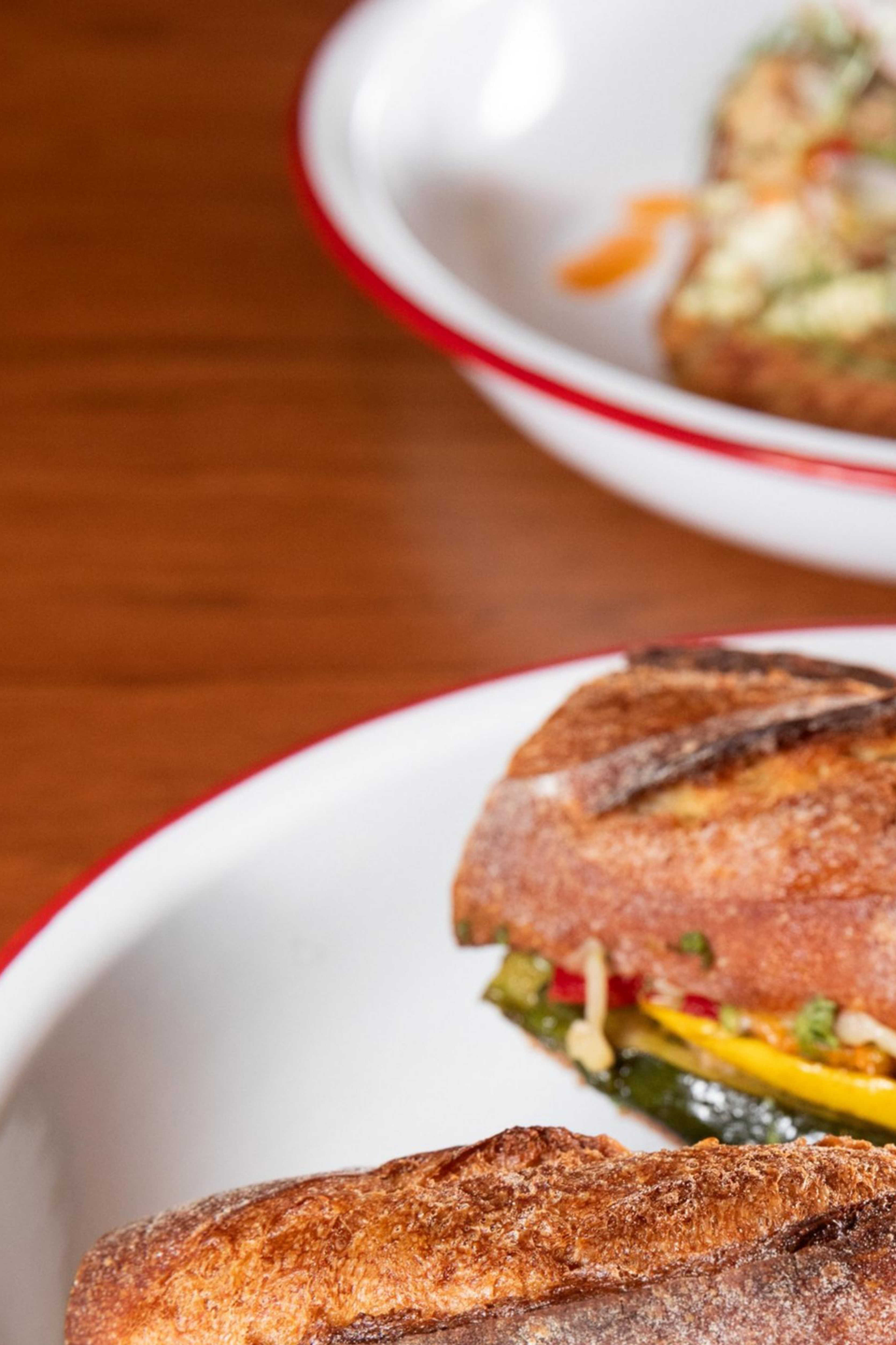 The image shows a crusty sandwich with roasted vegetables on a white plate with a red rim. In the background, there's an open-faced sandwich on a similar plate.