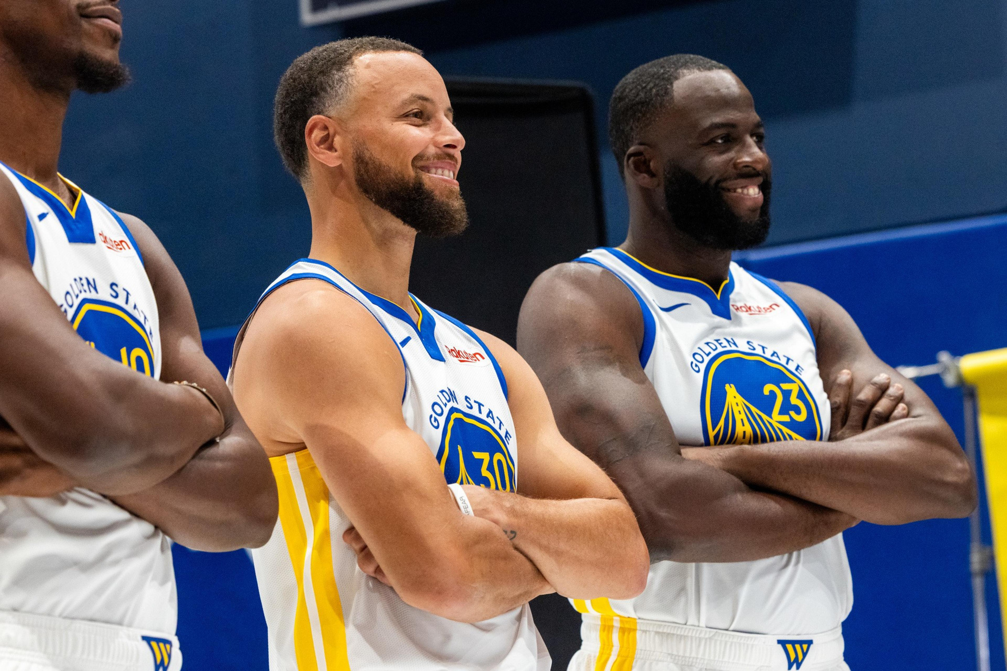 Three basketball players in Golden State Warriors uniforms stand side by side with arms crossed, smiling and looking off to the side.