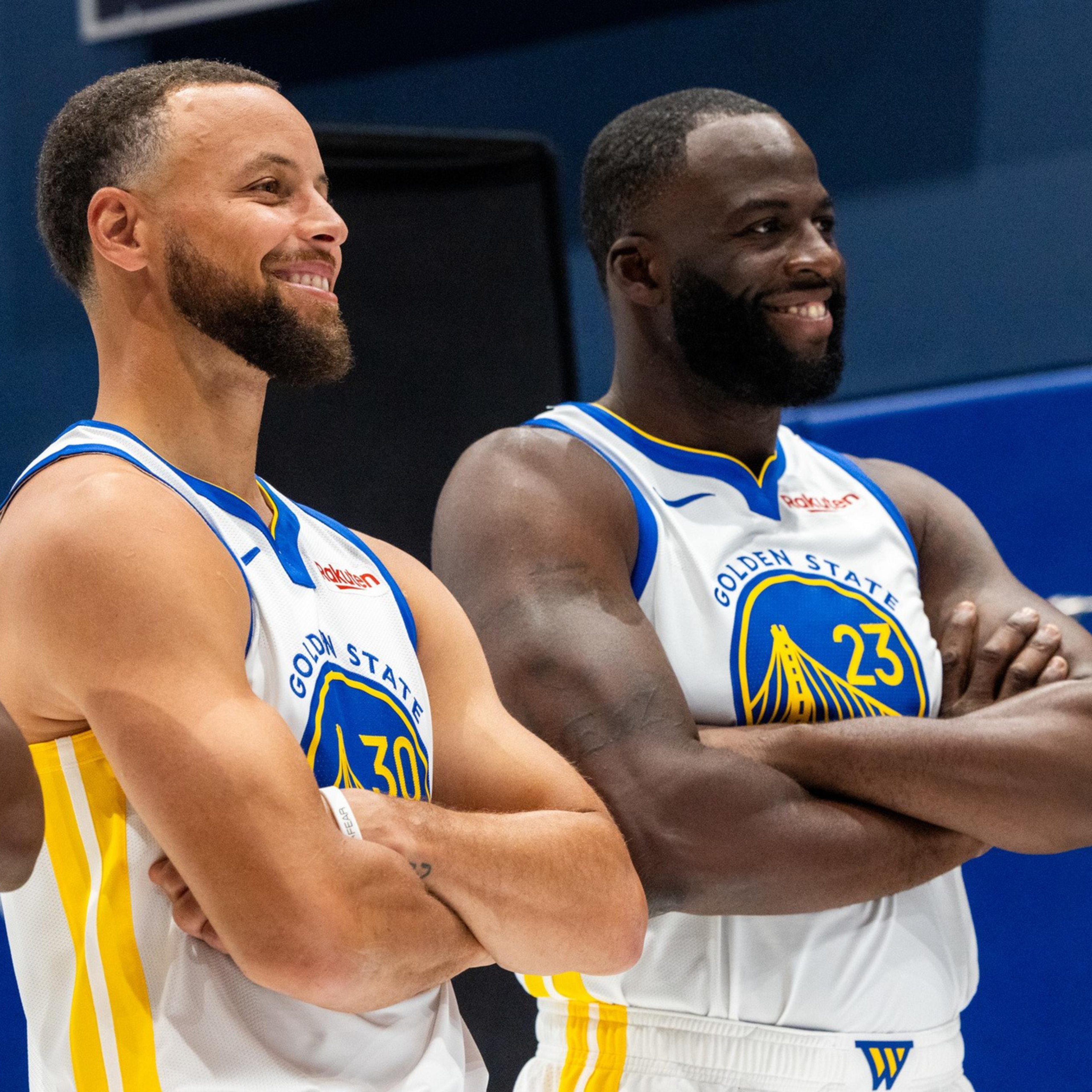Three basketball players in Golden State Warriors uniforms stand side by side with arms crossed, smiling and looking off to the side.
