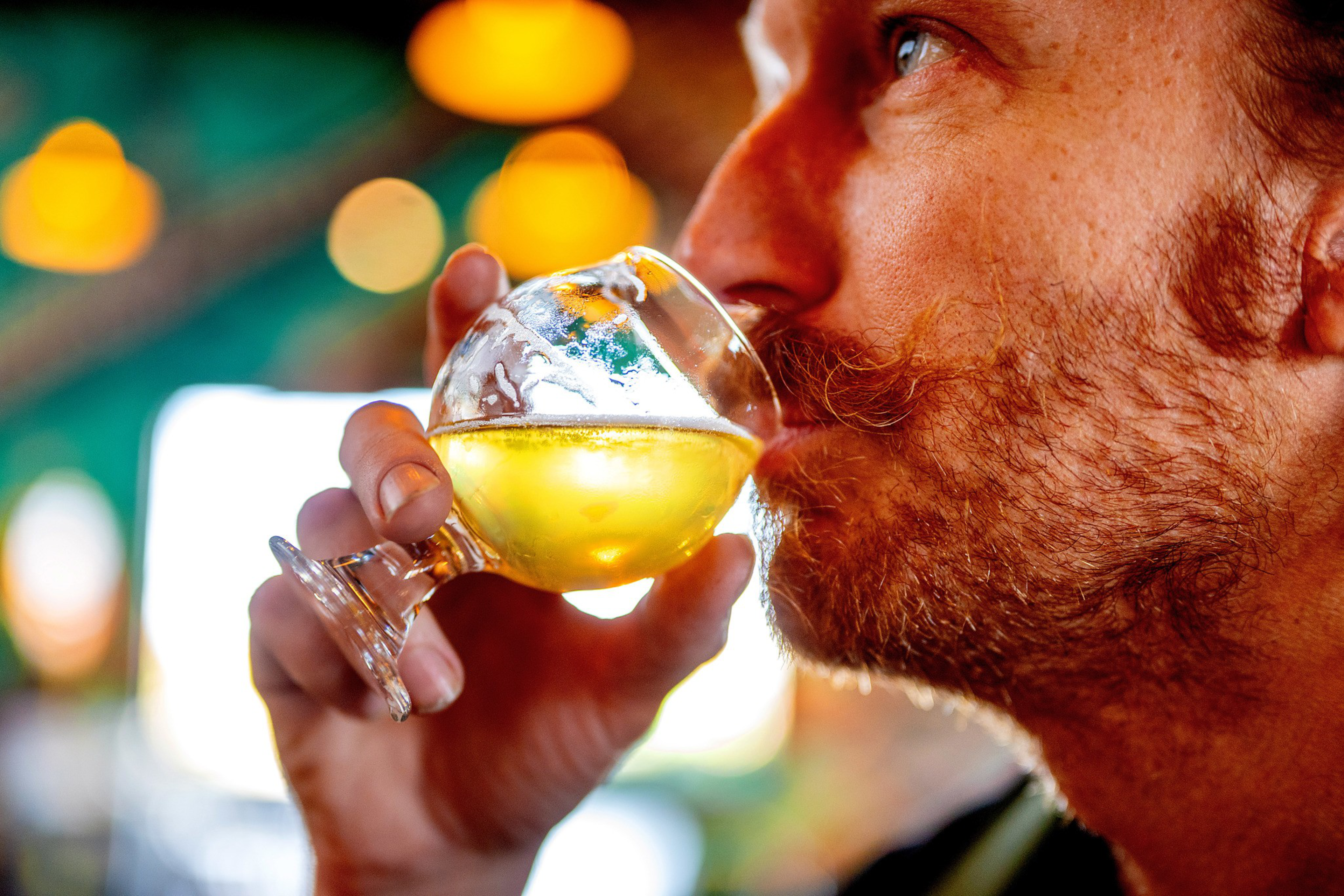 A man with a beard and mustache sips a golden-colored drink from a round glass, with soft, warm lights blurred in the background.
