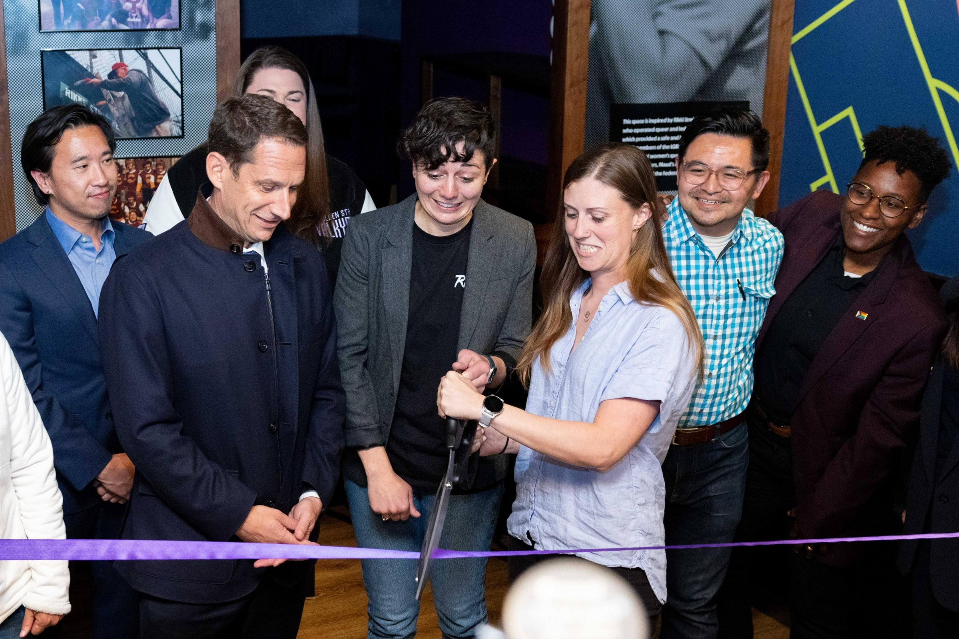 A group of people gather as a woman cuts a purple ribbon with large scissors, marking a ceremonial event. They all appear cheerful and engaged.