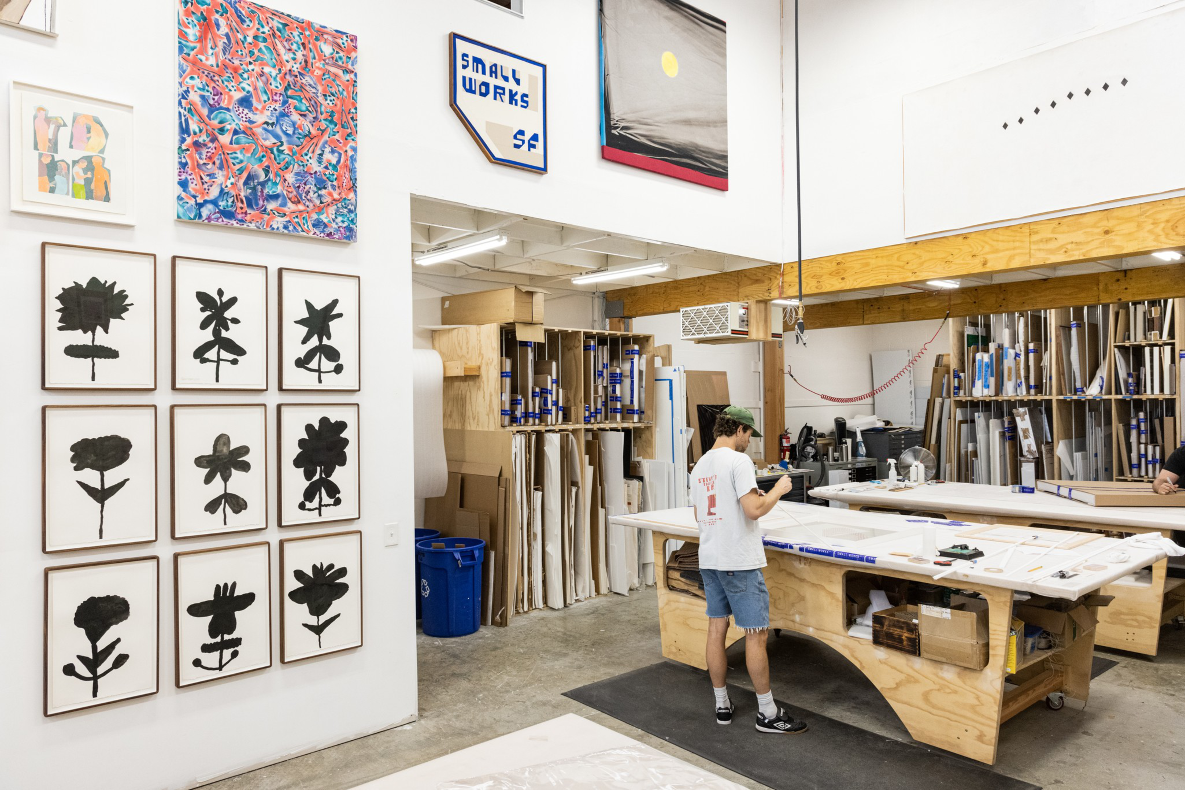 A person wearing a white T-shirt and denim shorts stands at a large wooden worktable in an art studio with framed black flower prints on the wall.
