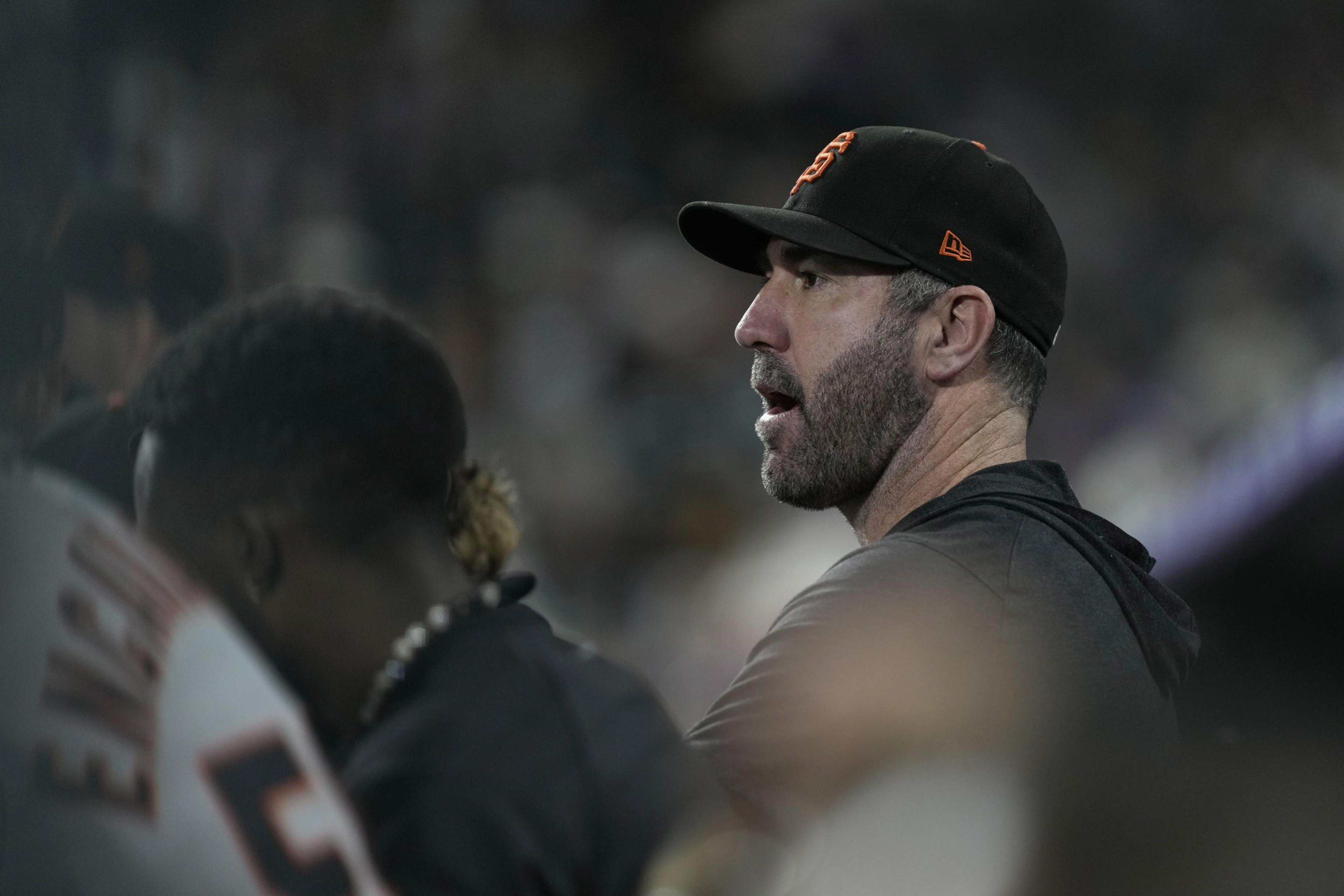 A man wearing a black baseball cap with an orange logo and a black shirt is speaking, while another person wearing a white jersey with the number 5 is beside him.