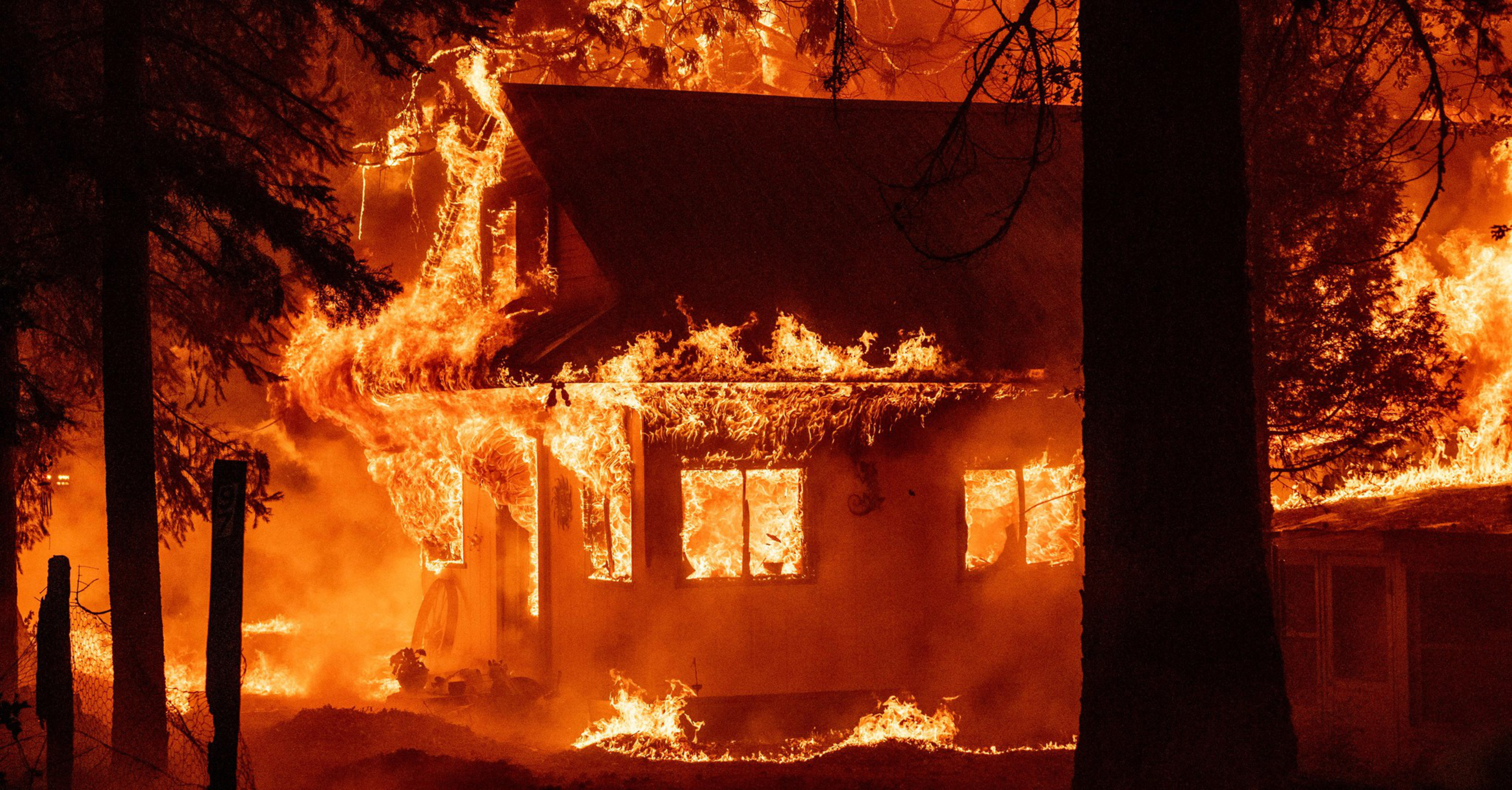 A house is engulfed in intense flames at night, surrounded by tall trees, with a bright orange glow illuminating the scene.