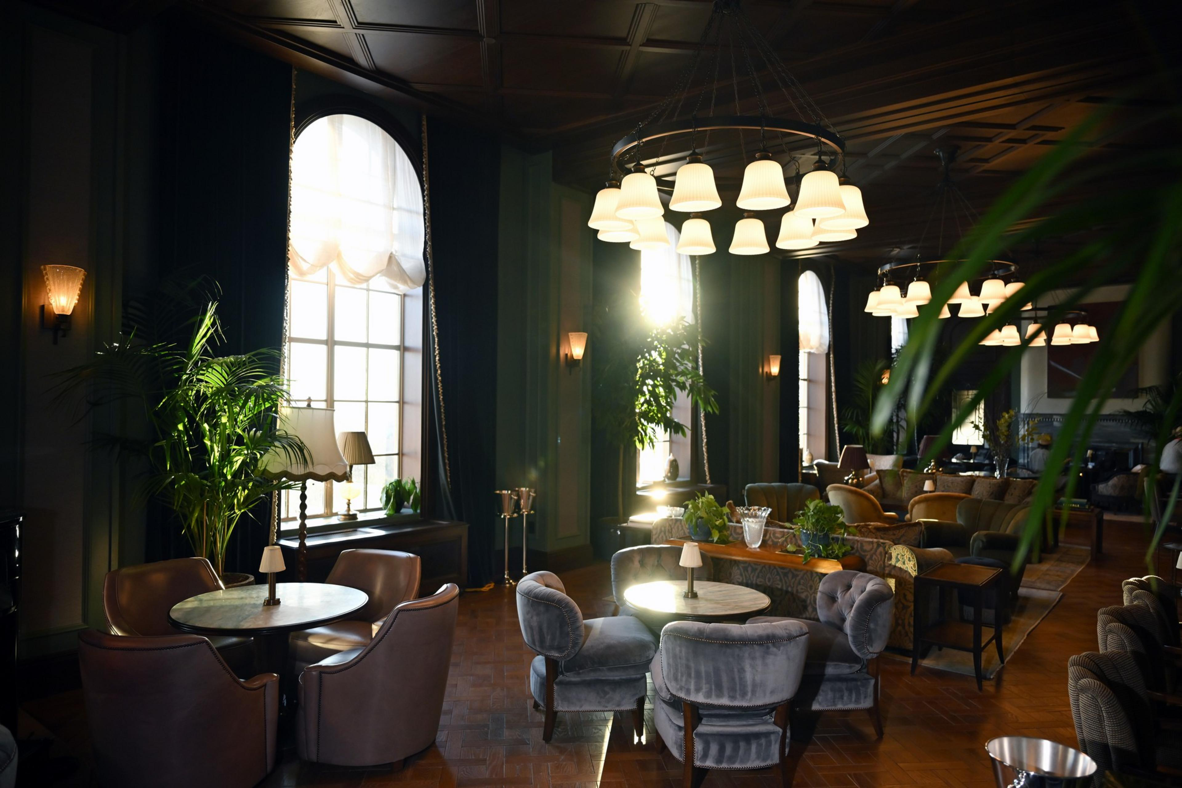 The image shows a cozy, upscale lounge with round tables, plush chairs, large windows with curtains, and warm lighting. Plants decorate the space.