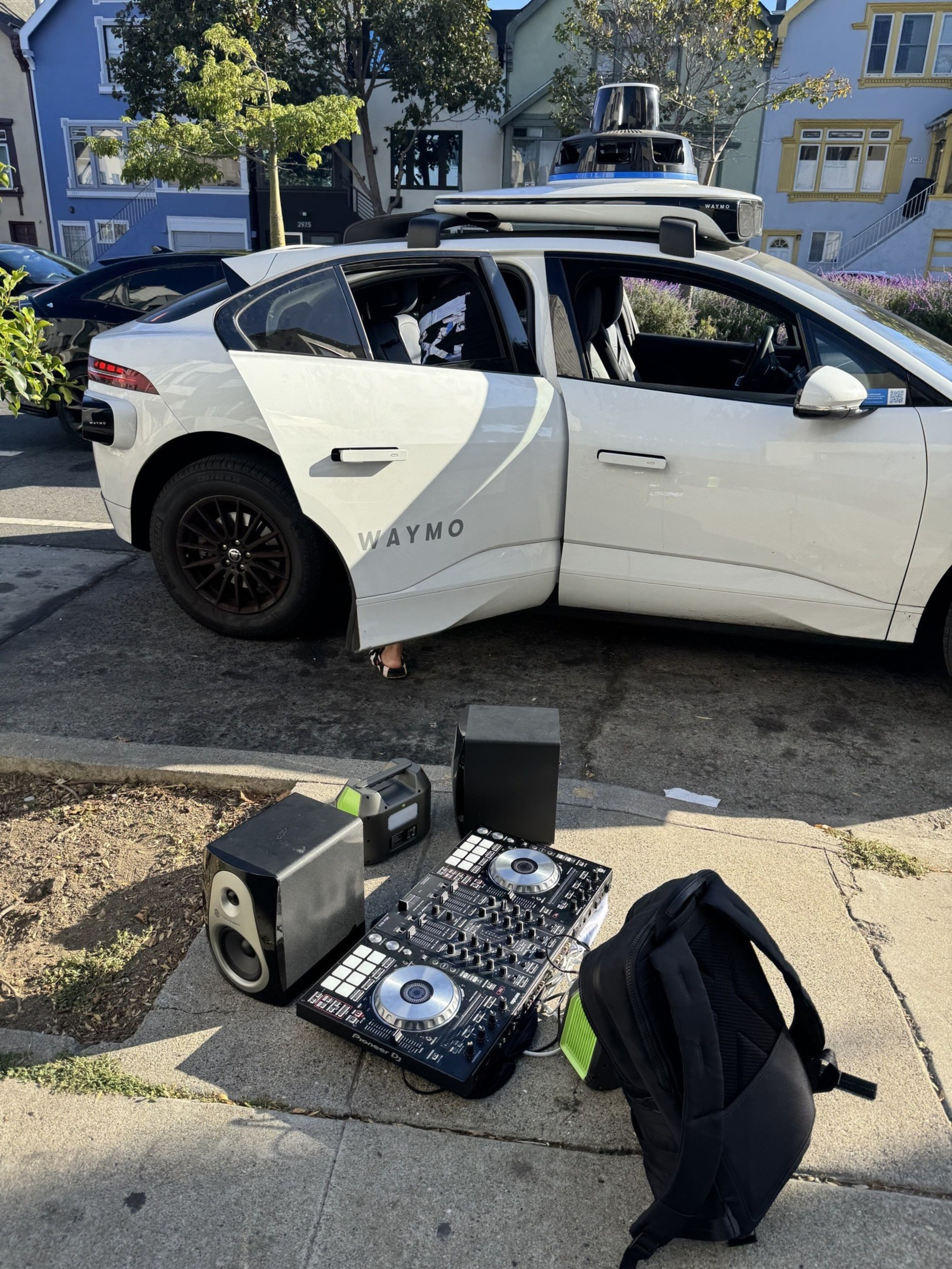 A white Waymo self-driving car is parked on a street. Nearby, there's DJ equipment on the sidewalk with speakers and a backpack.