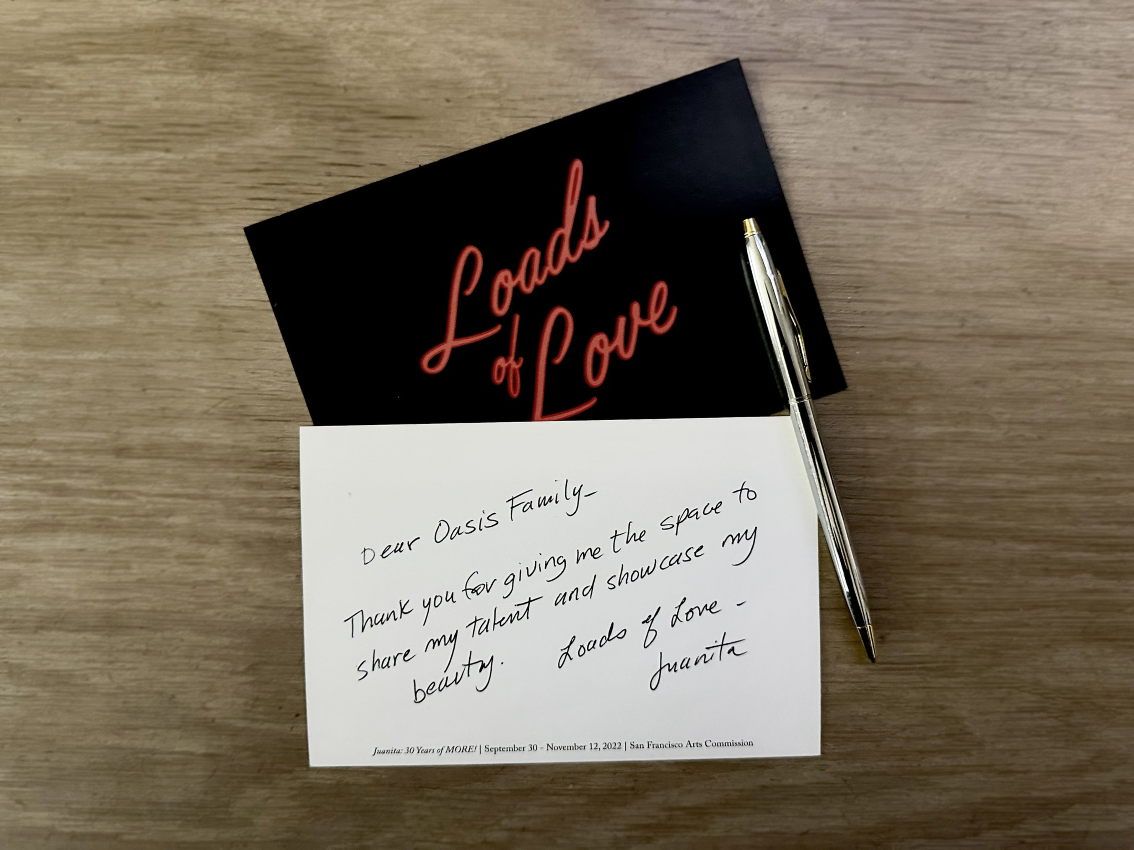 A handwritten thank-you note on white paper and a black card with "Loads of Love" in red cursive lie on a wooden surface, next to a silver pen.