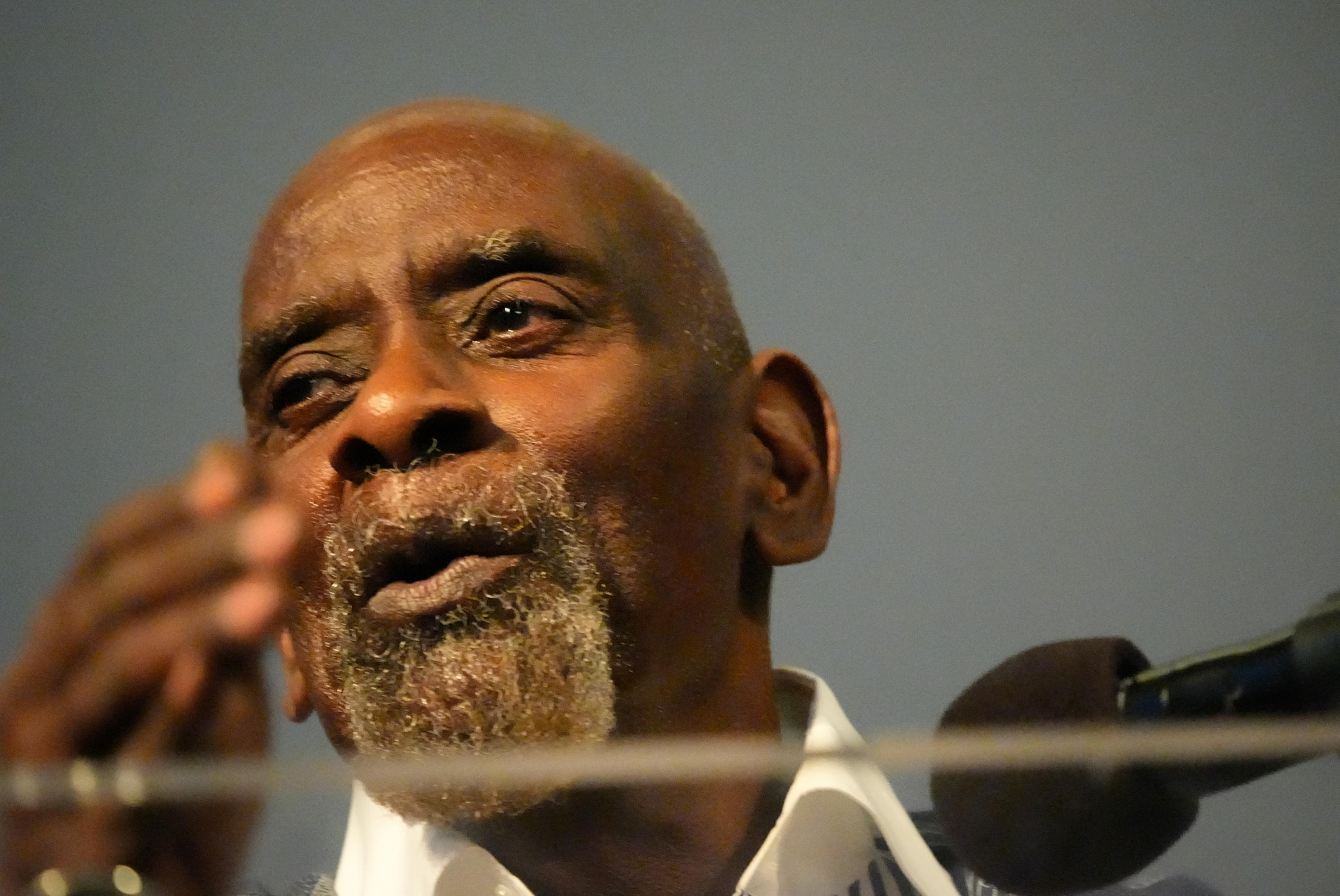 An older man with a grey beard speaking into a microphone, appearing contemplative or in mid-speech.