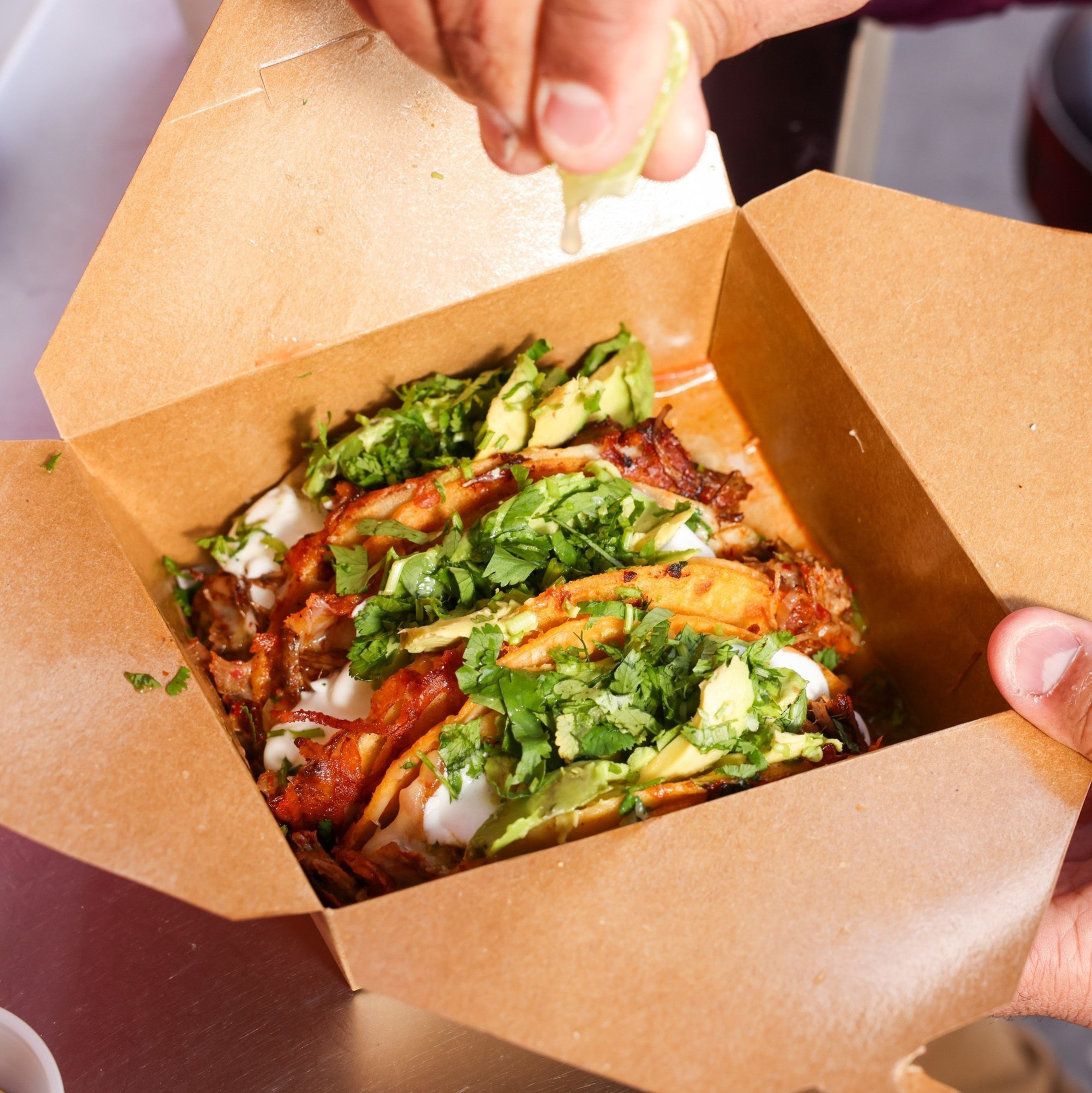 Three tacos with cilantro, avocado, and cream are in an open brown box. A hand squeezes lime juice over them.