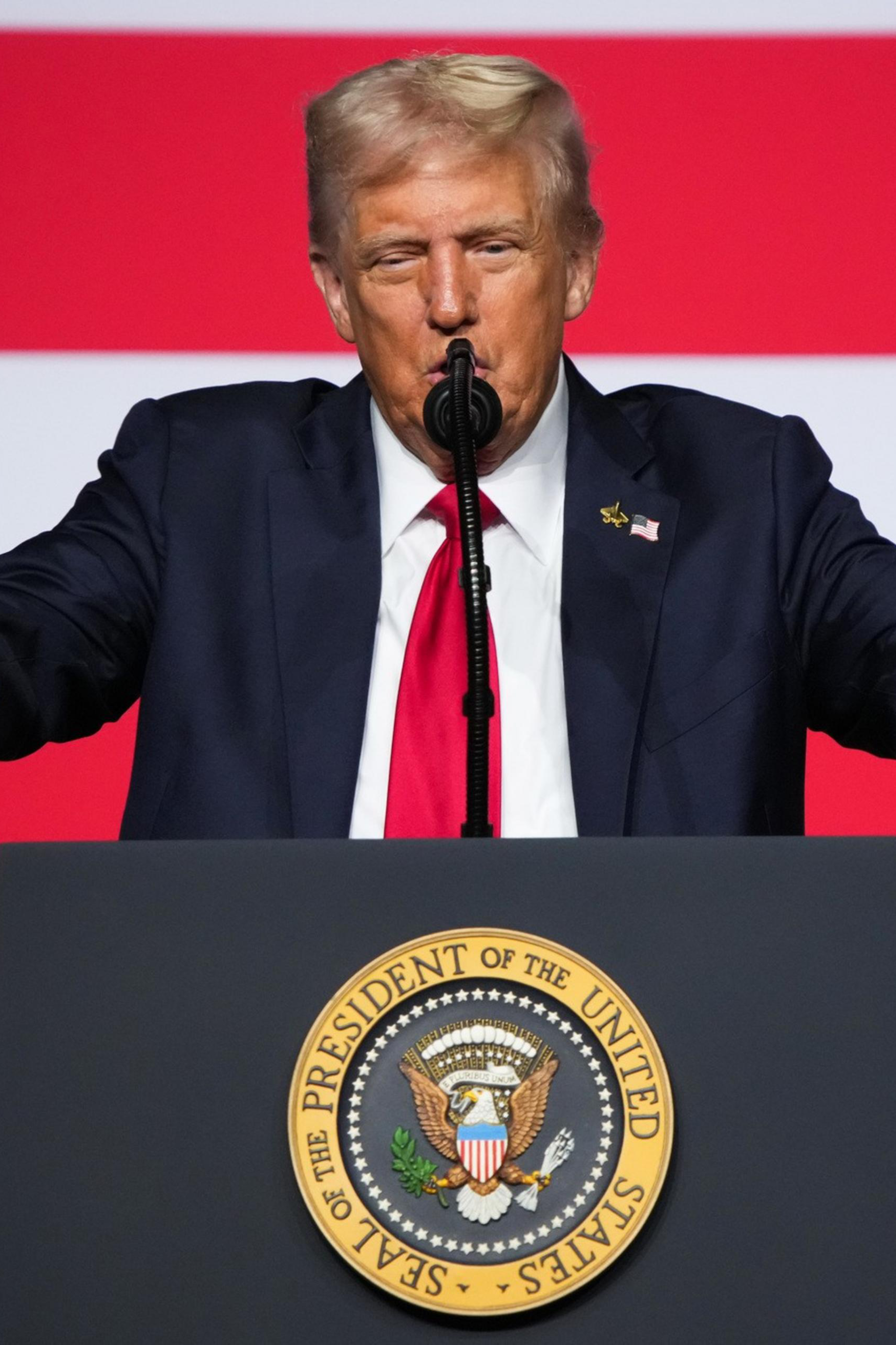 A man in a suit with a red tie stands behind a podium bearing the presidential seal, speaking in front of a large American flag.