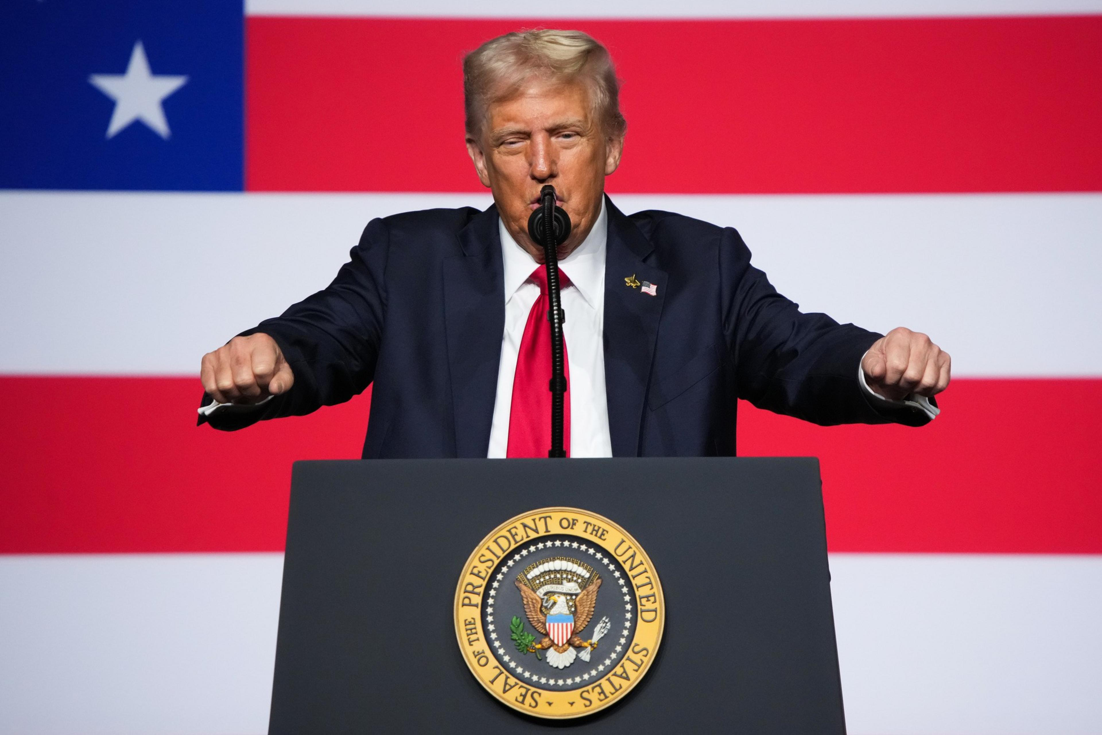 A man in a suit with a red tie stands behind a podium bearing the presidential seal, speaking in front of a large American flag.