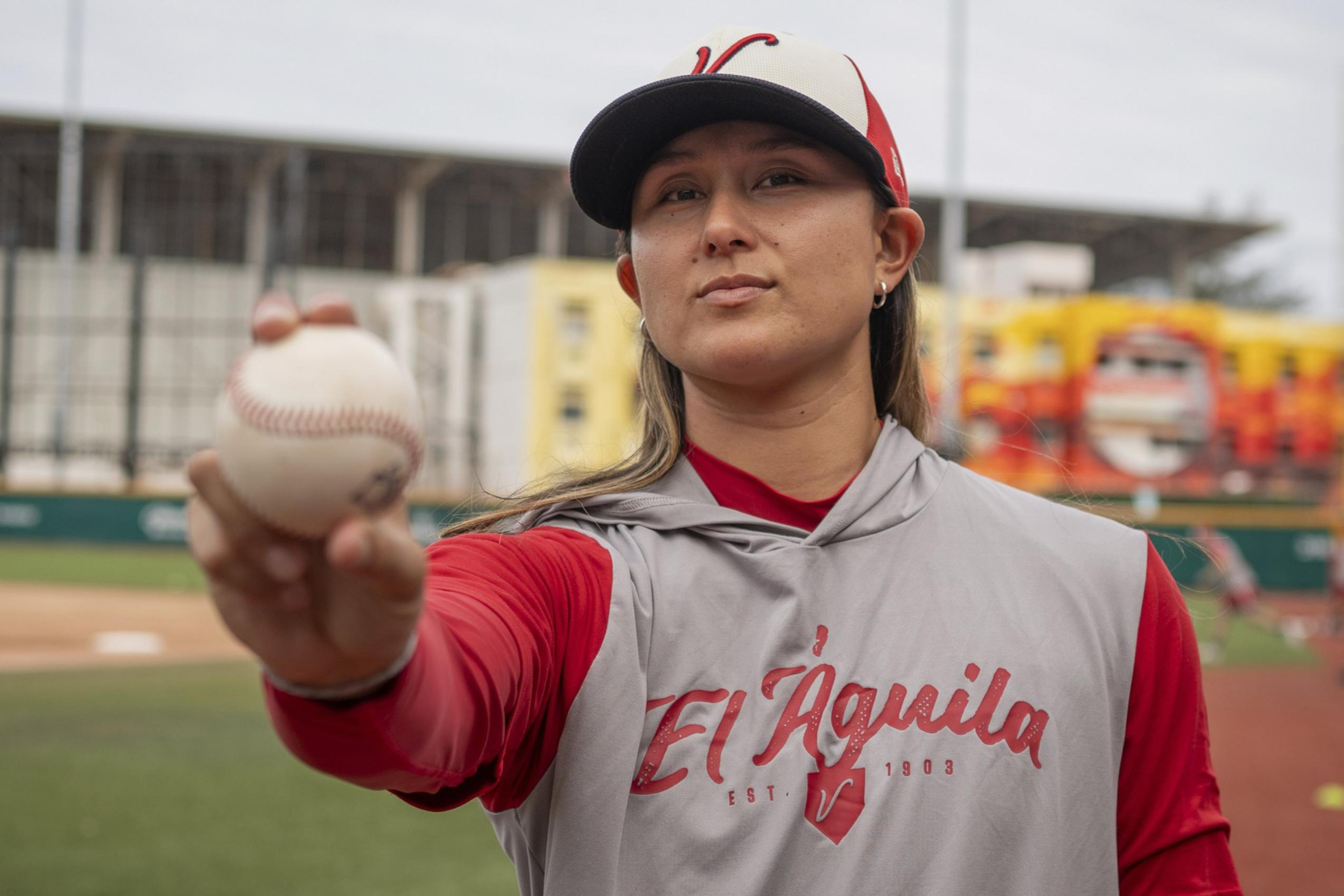 A baseball player wearing a red and gray uniform with “El Águila” on the front holds a baseball toward the camera on a field.