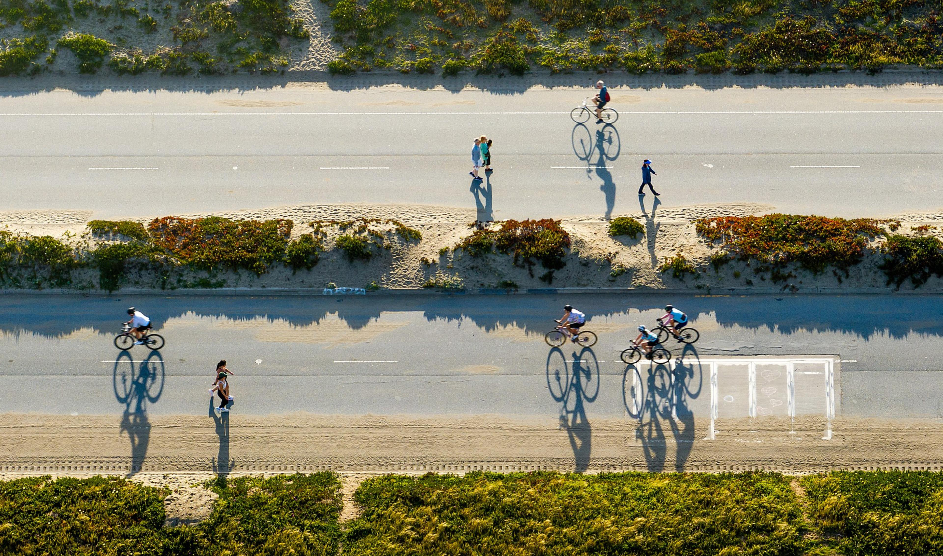 An aerial view shows people biking and walking on parallel paths, with shadows cast on the ground. Greenery lines the upper and lower edges of the image.