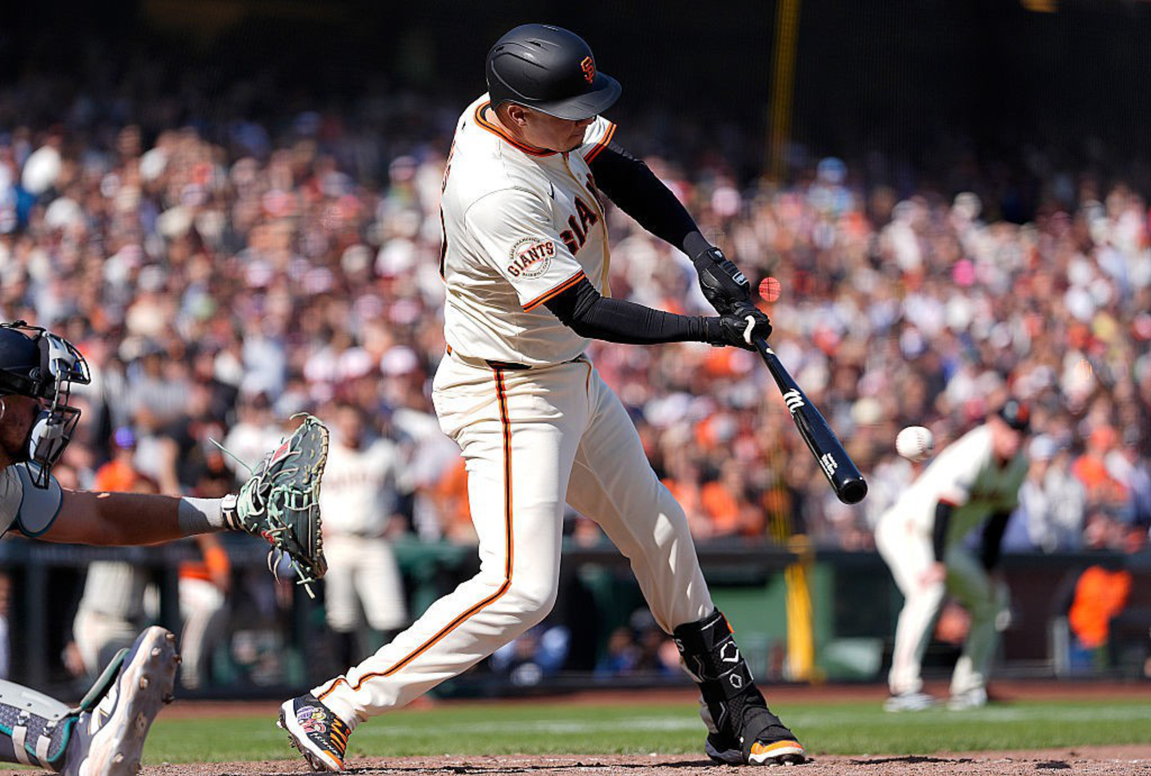 A baseball player in a Giants uniform is mid-swing during a game, with a catcher and umpire nearby, and a crowded stadium in the background.