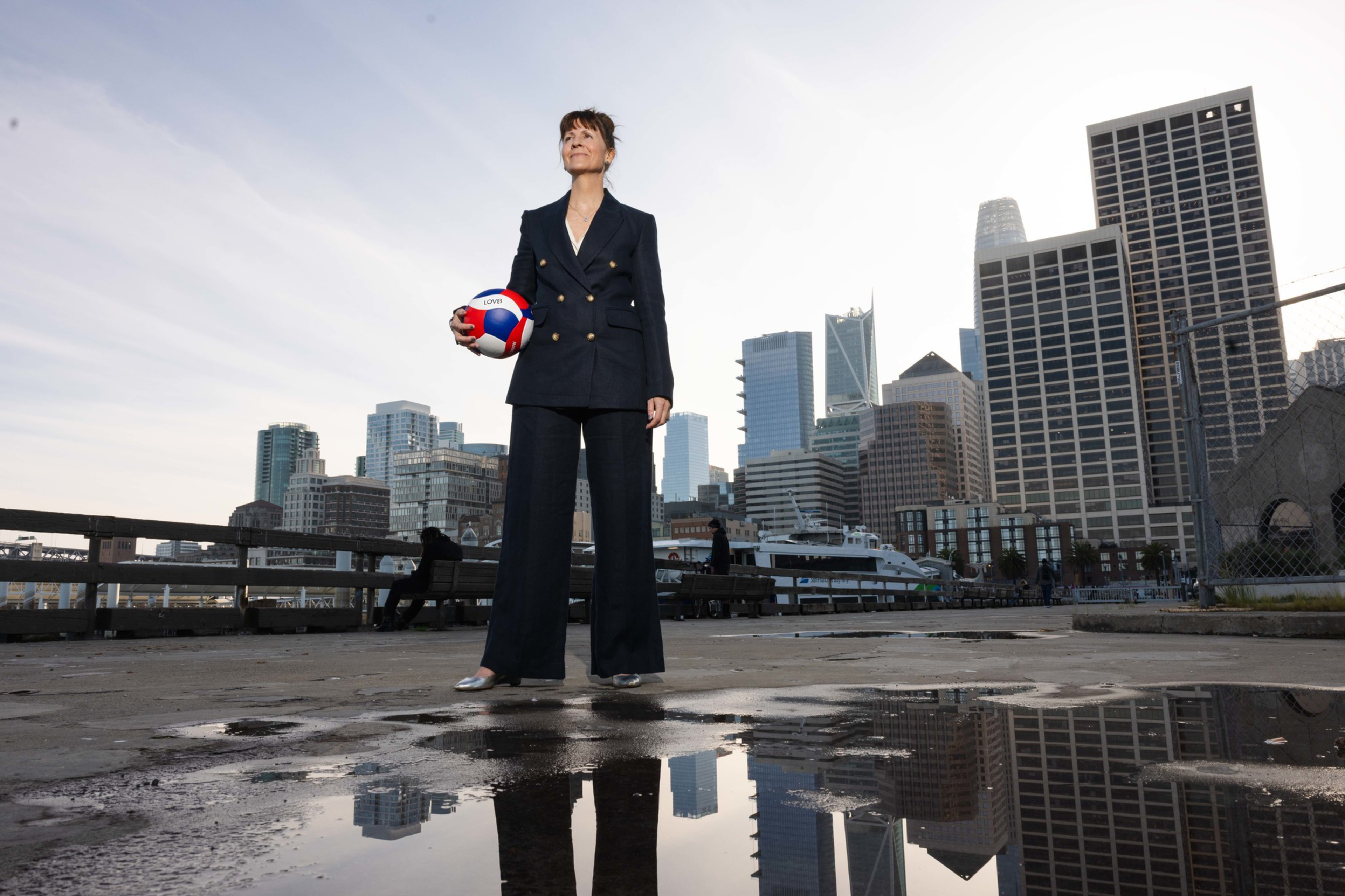 A woman in a black suit holds a volleyball while standing on a wet dock with city skyscrapers reflected in a puddle beneath her.