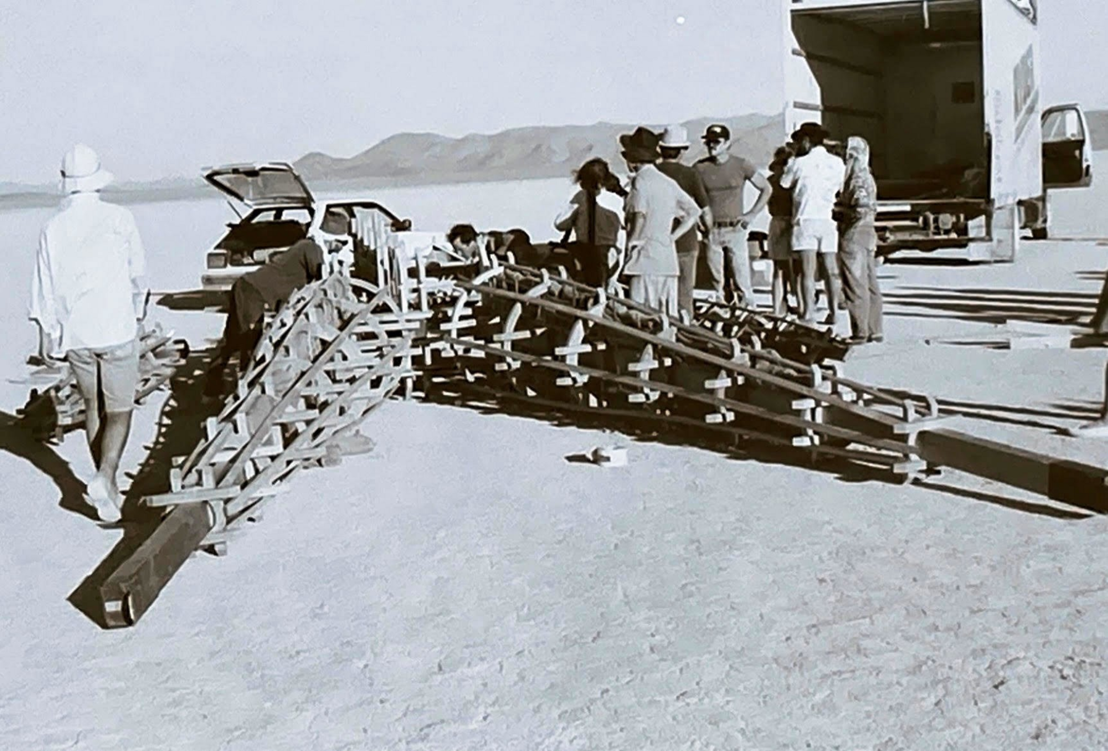A group of people stands and works around a large, wooden, triangular framework structure on a flat, desert-like surface with vehicles parked nearby.