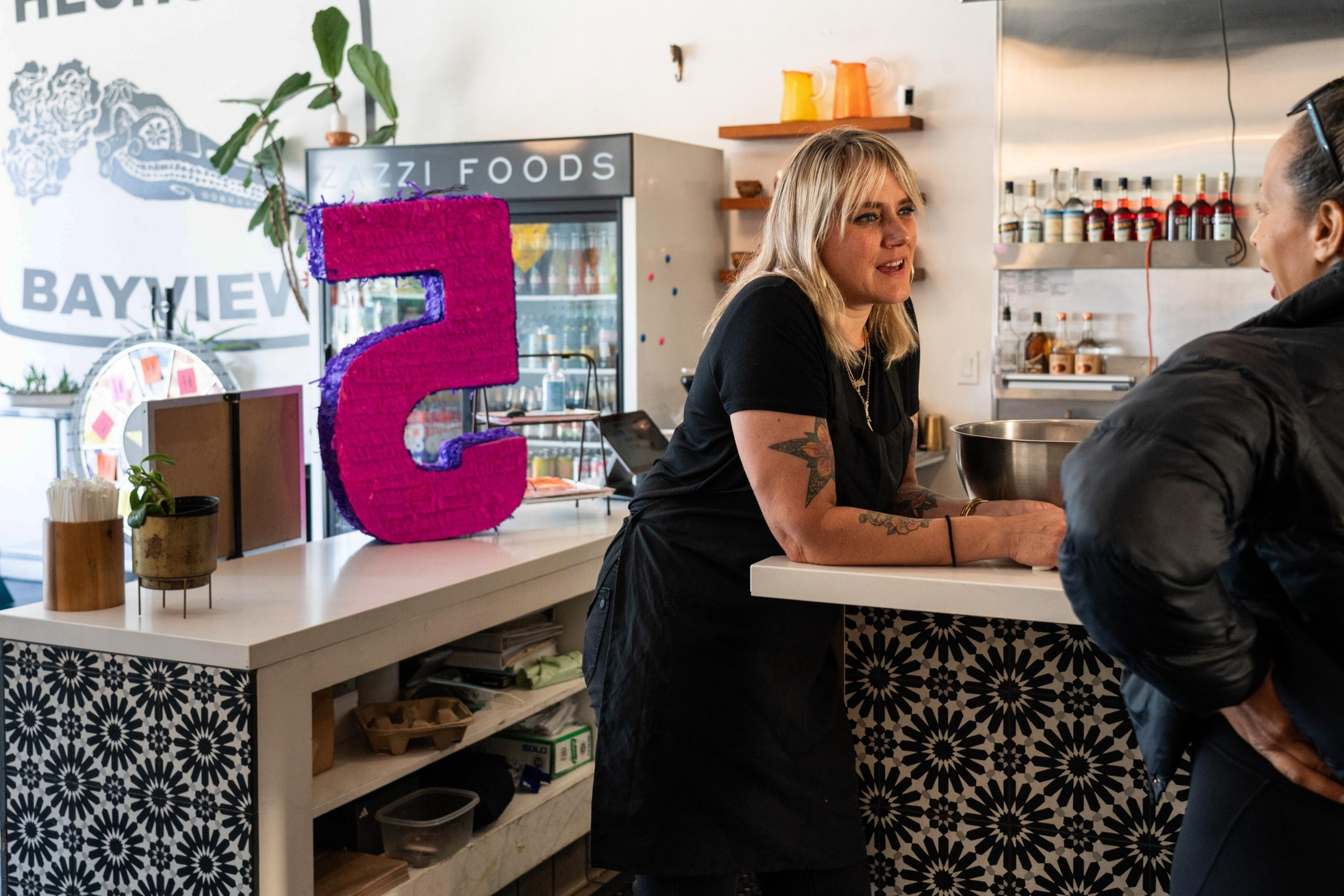 A woman converses with a customer across a café counter, with a vibrant pink piñata in the shape of the number 2 in the foreground.