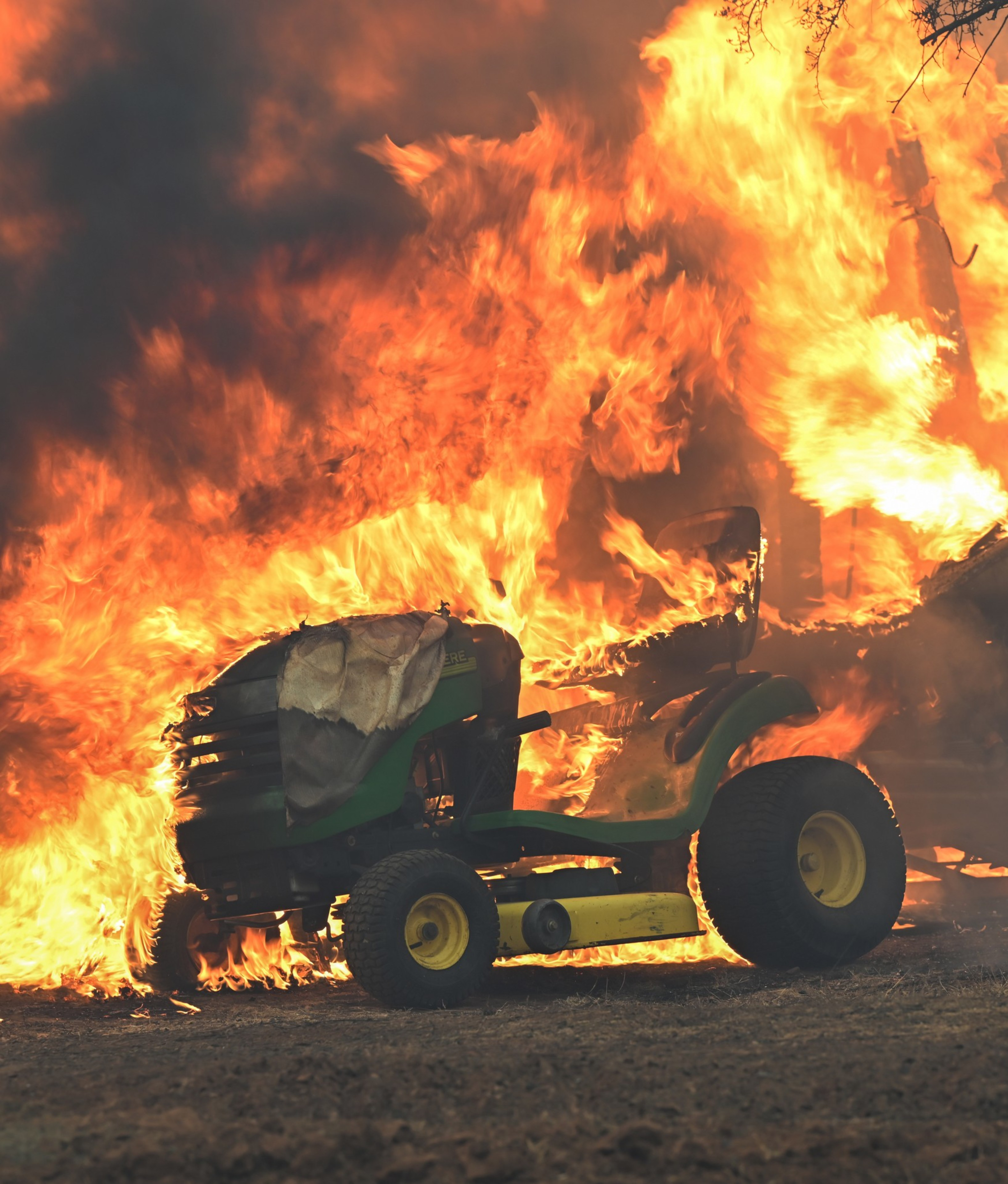 A small green tractor is engulfed in intense flames, with thick smoke rising above. The fire is consuming the tractor and spreading to nearby objects.