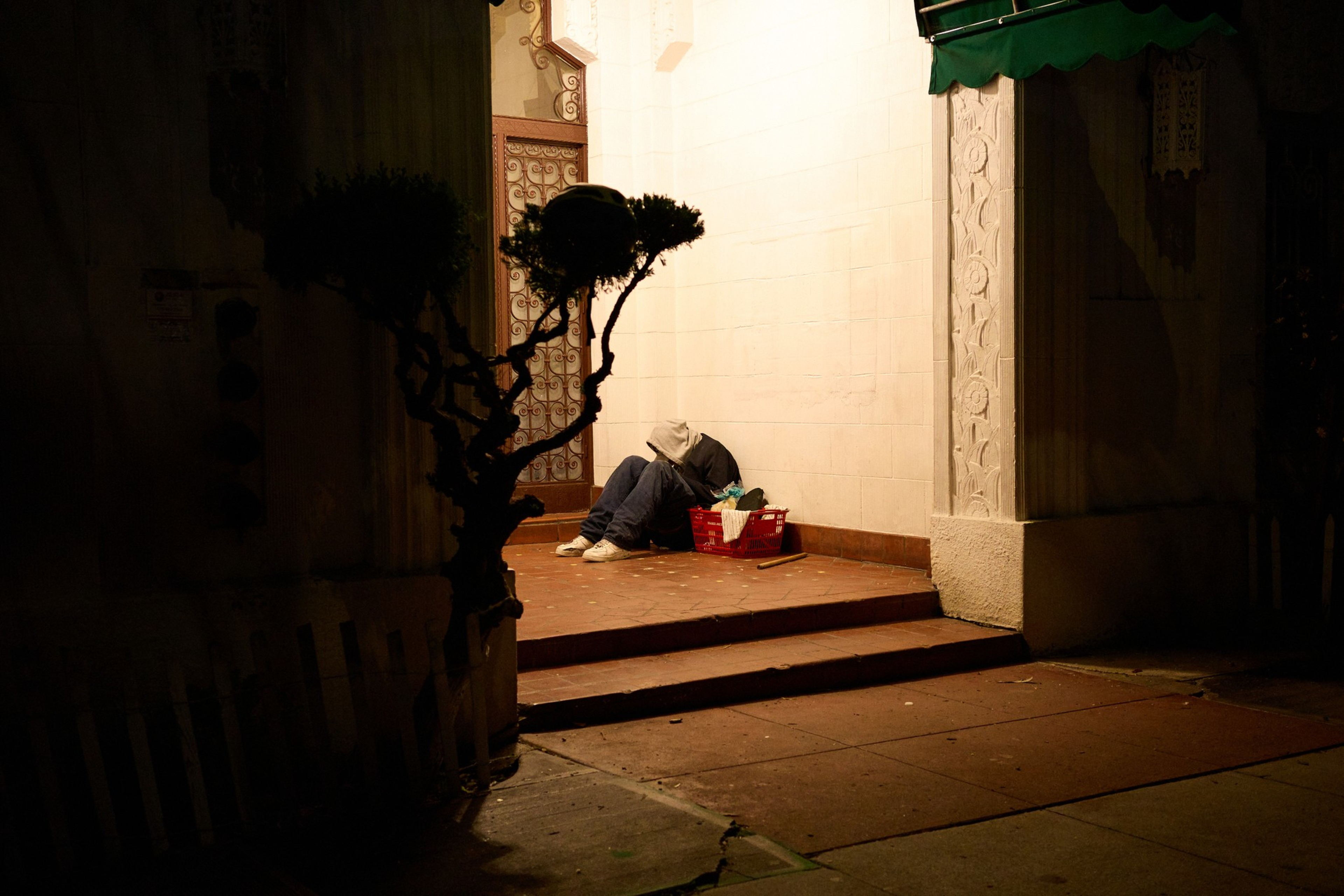 A person in a hoodie sits on steps in a dimly lit alcove, next to a red basket filled with belongings. A small tree casts shadows nearby.