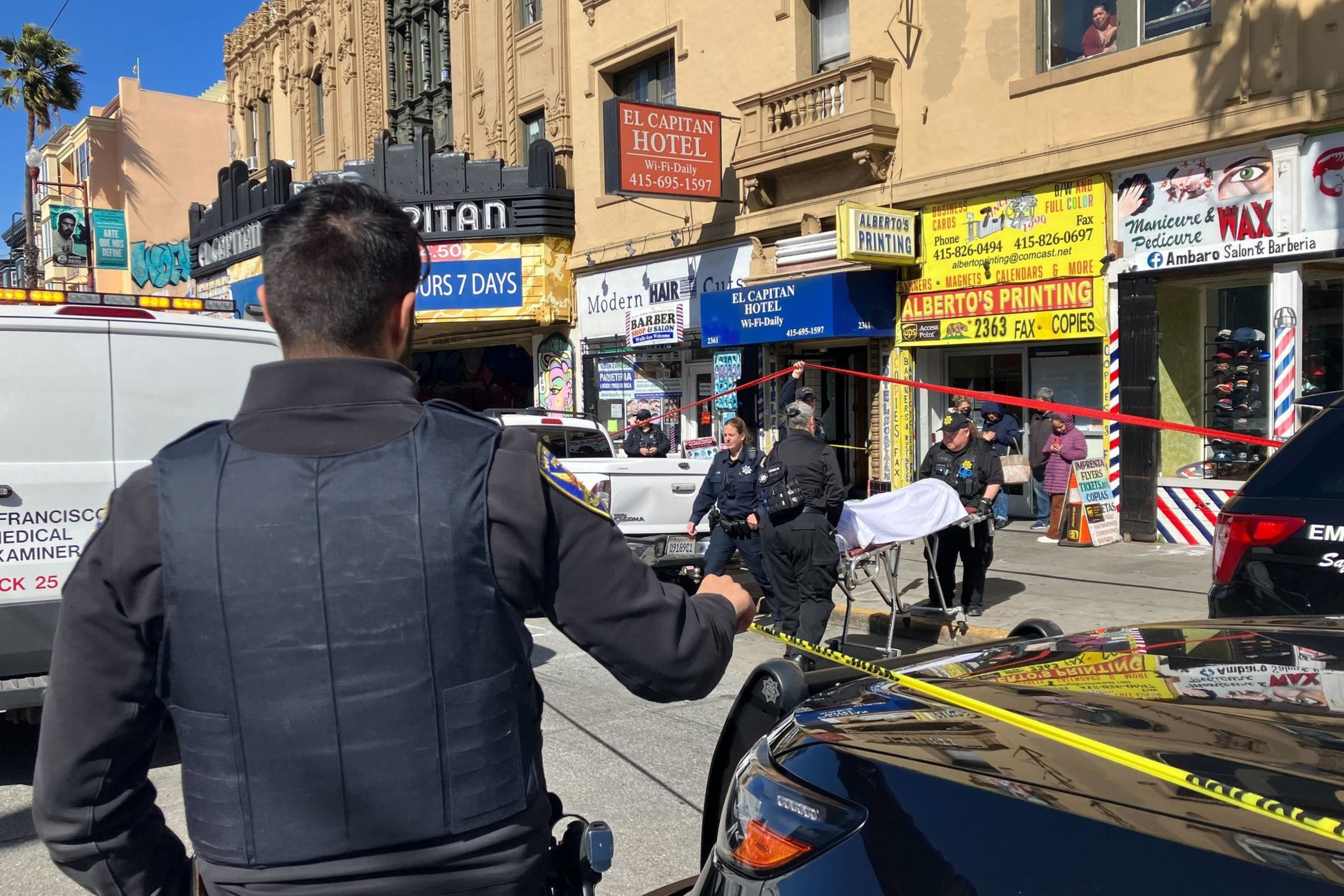 A police officer observes a cordoned-off urban scene with emergency personnel and onlookers.