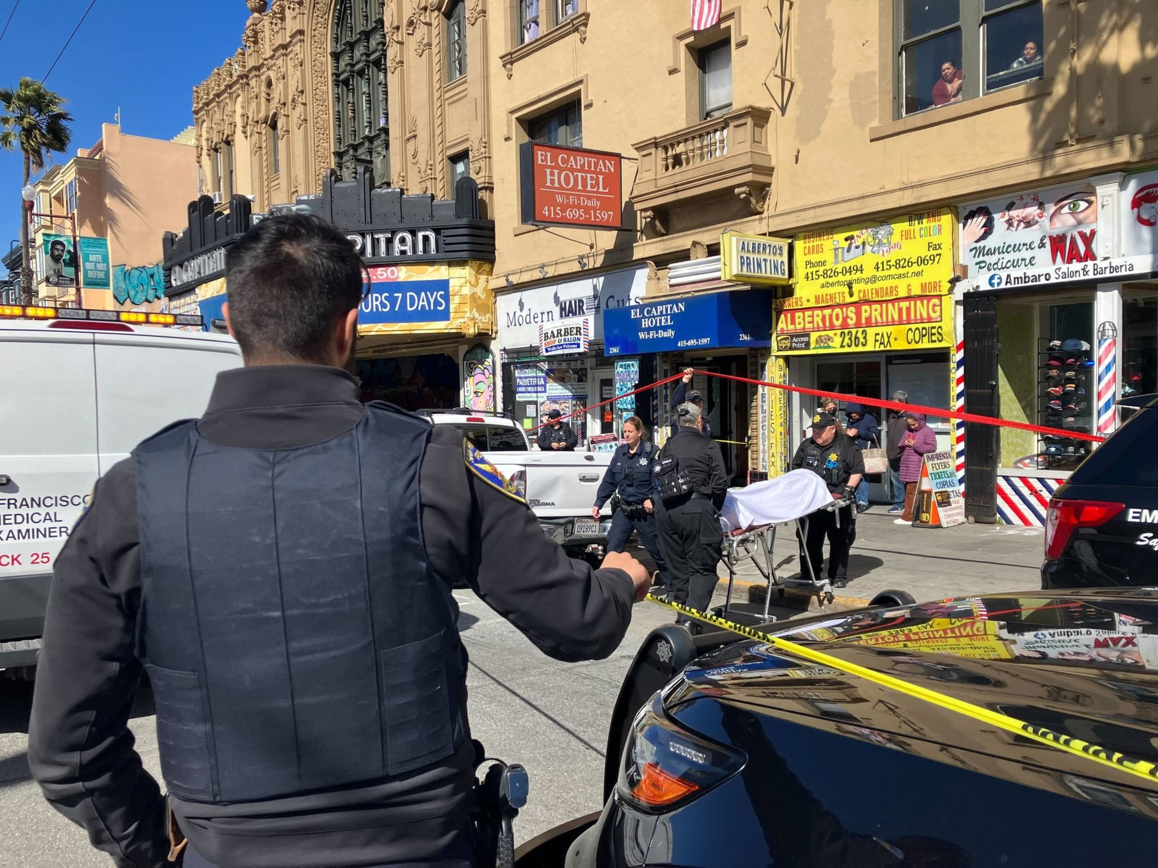 A police officer observes a cordoned-off urban scene with emergency personnel and onlookers.