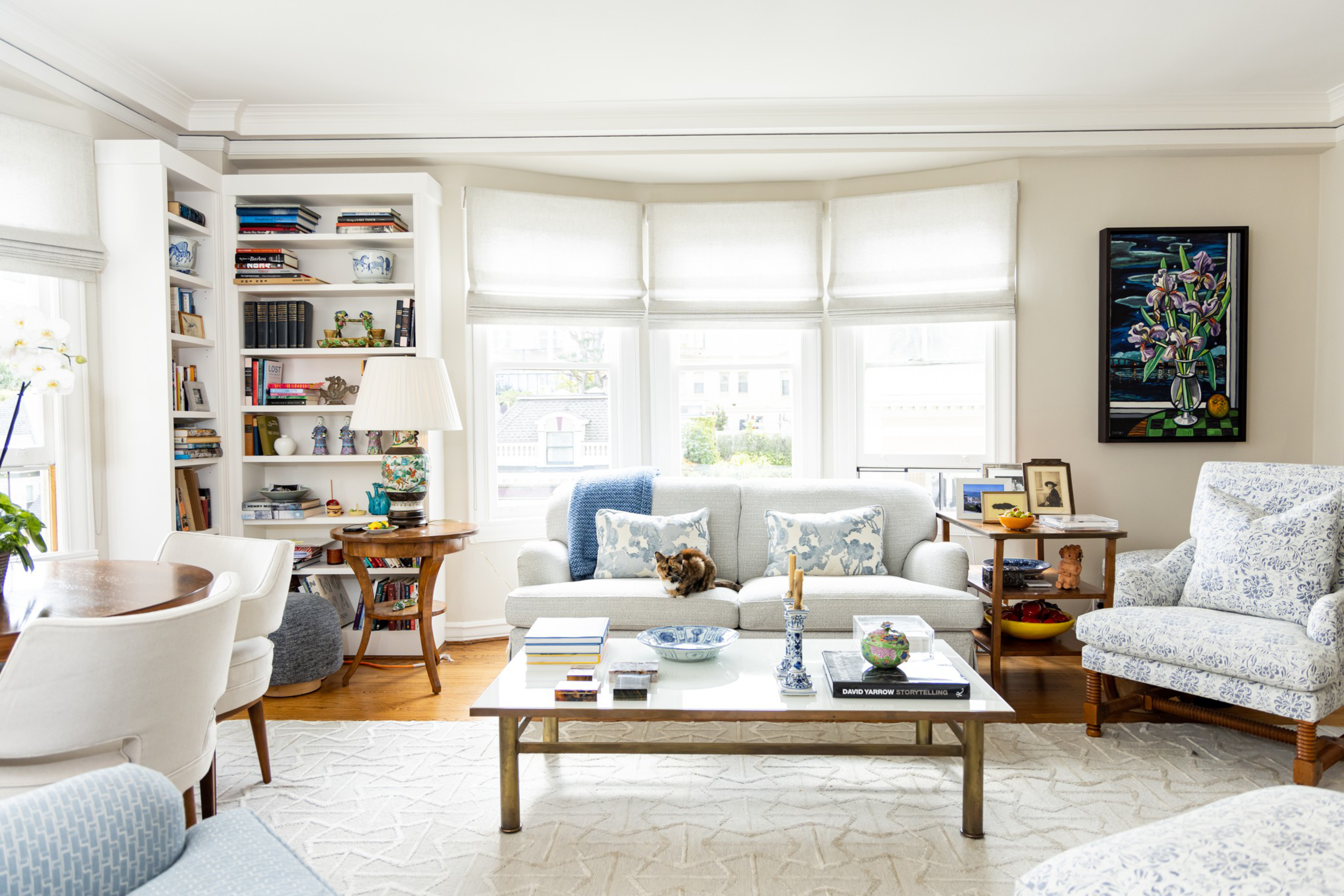 A bright living room with white walls and furniture, a central coffee table with books and decor, a sofa with pillows and a cat, flanked by armchairs and side tables.
