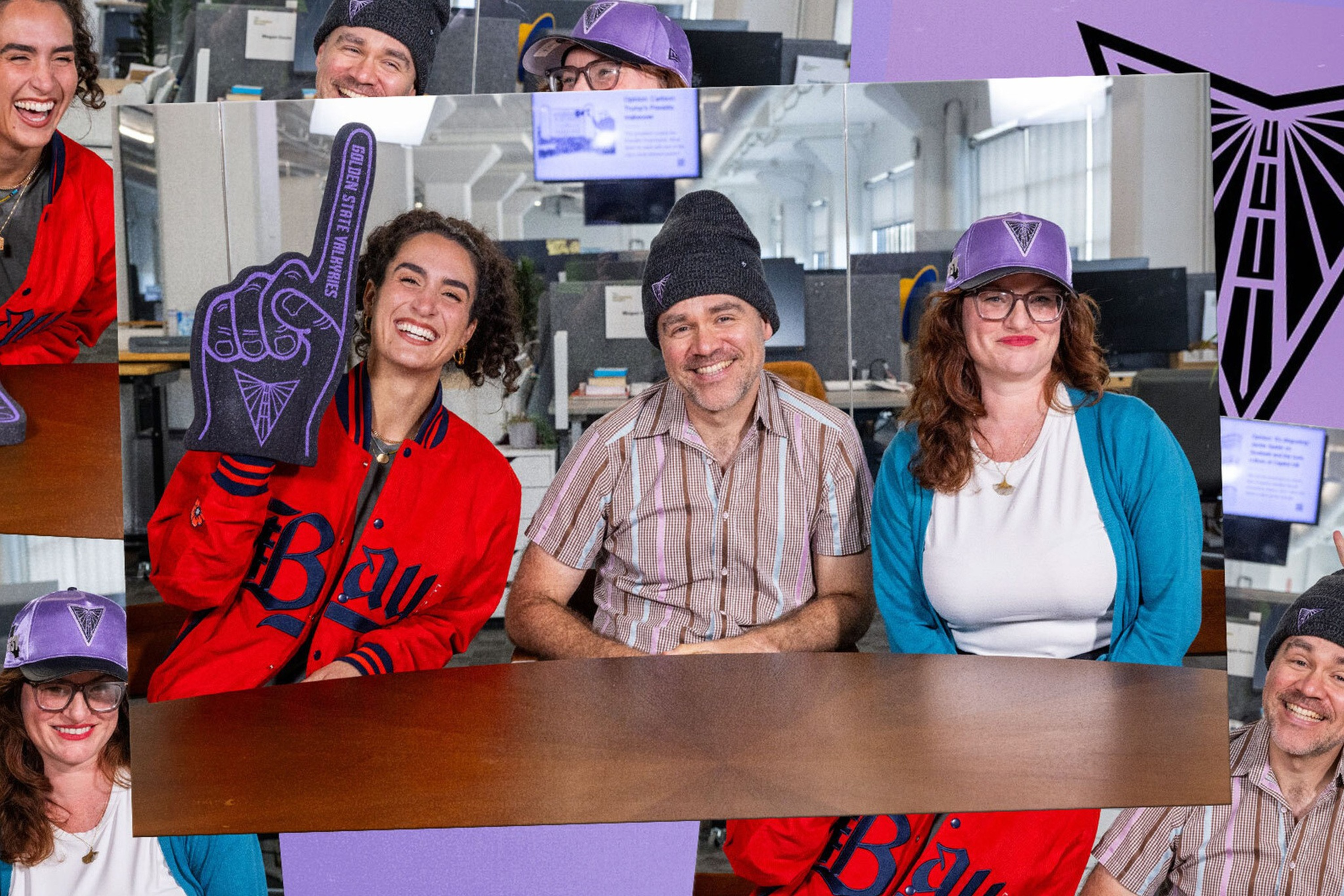 Three people sit at a table in an office: a woman in a red jacket holds a foam finger, a man in a beanie smiles, and a woman in glasses and a purple cap looks on.
