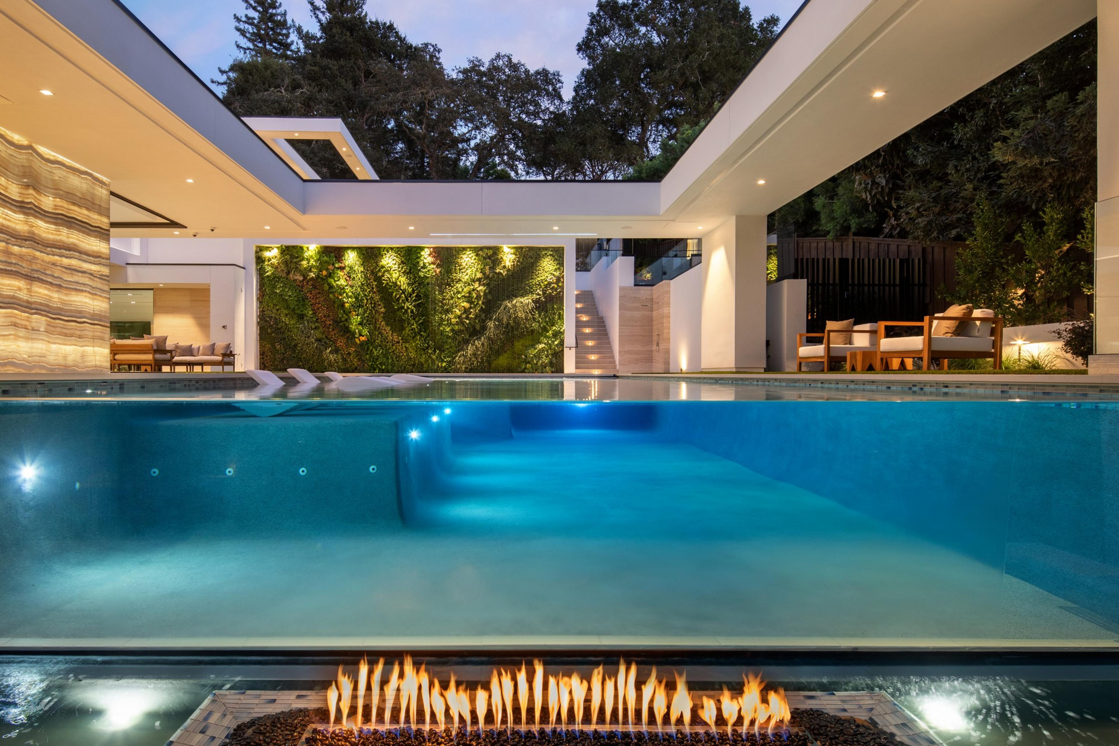 A modern indoor-outdoor space with a glass-sided pool, seating areas, vertical garden, and a sleek linear fire feature.